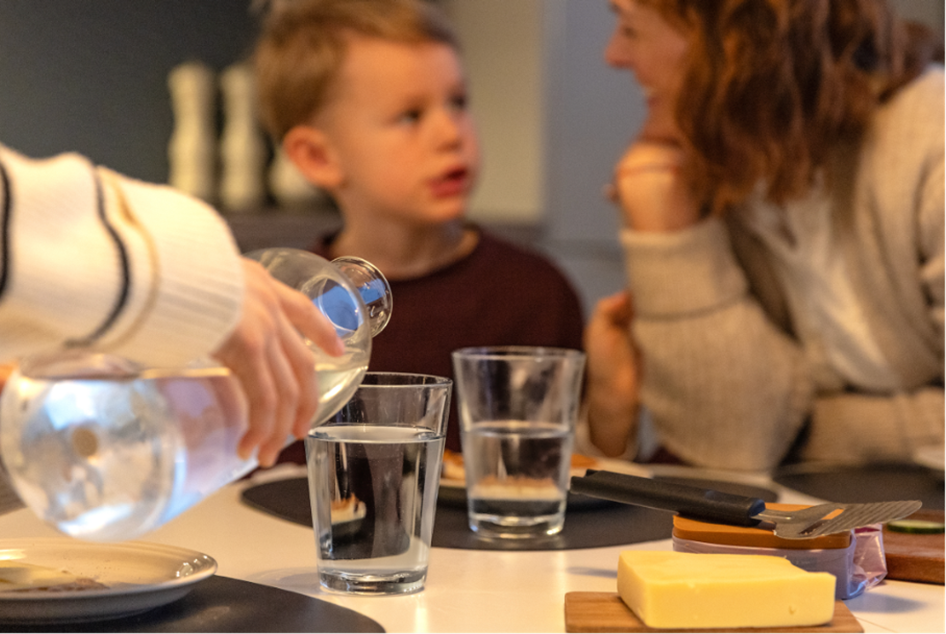 Familiemåltid der vann skjenkes i glass mens et barn og en voksen samtaler ved bordet med smør og bestikk synlig.