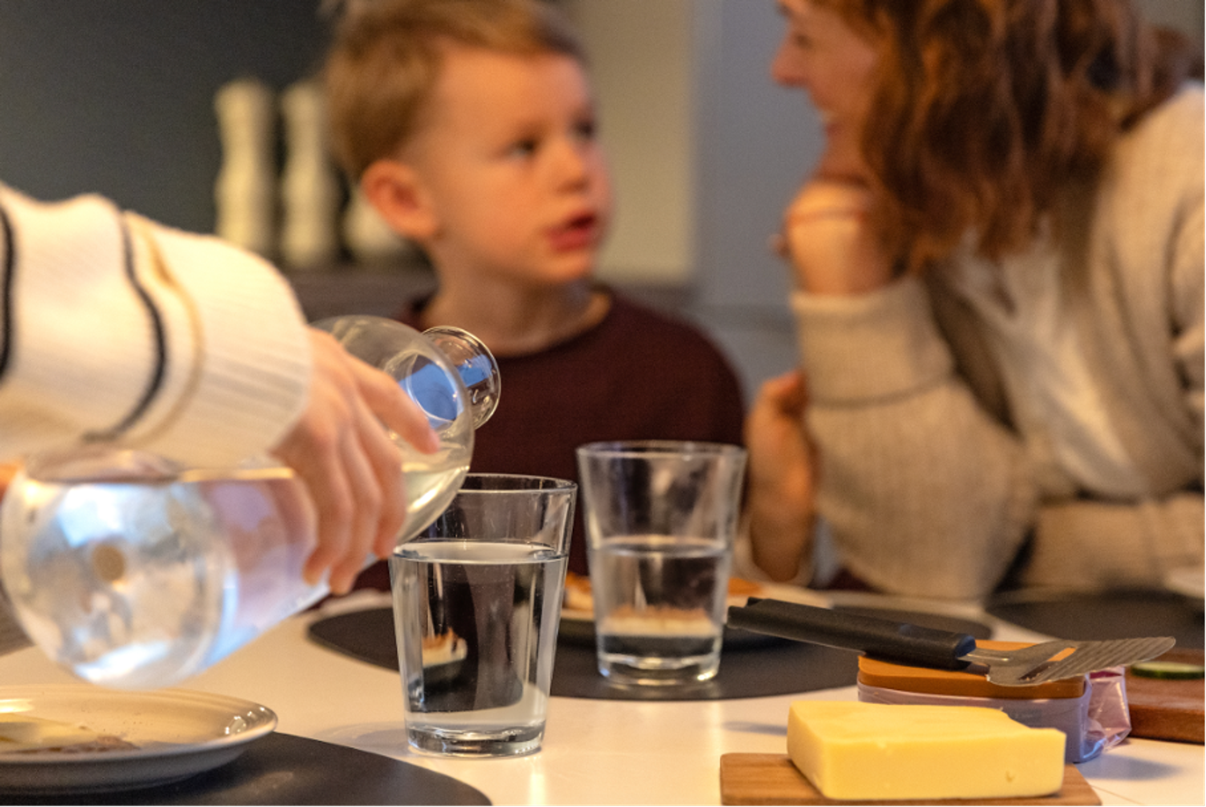 Familiemåltid der vann skjenkes i glass mens et barn og en voksen samtaler ved bordet med smør og bestikk synlig.