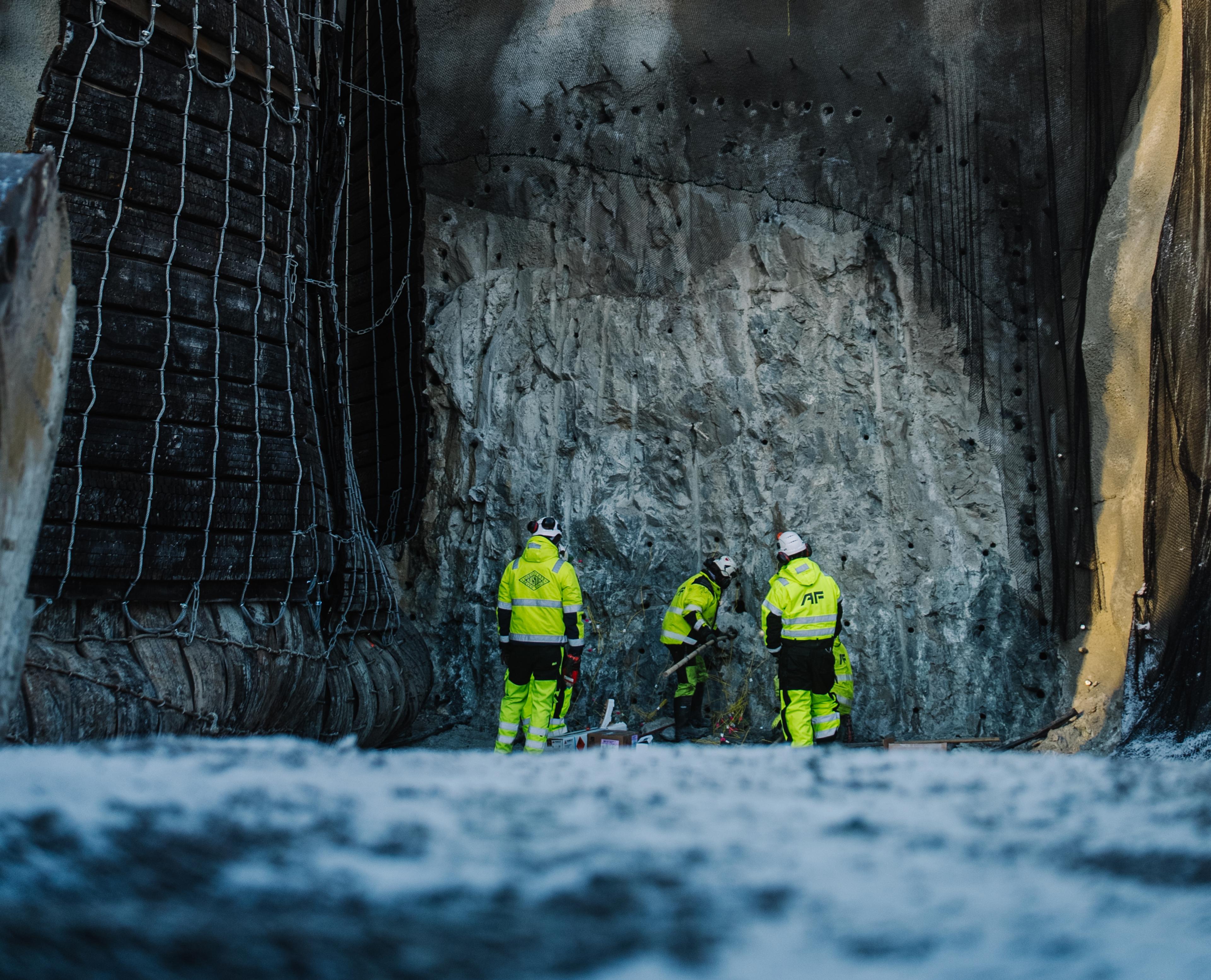Bildet viser et anleggsområde for tunnelbygging om vinteren. Tre anleggsarbeidere i gule refleksvester står ved en høy betongvegg som er sikret med nett og støttekonstruksjoner. Ovenfor anleggsområdet er det snødekte skråninger og nakne trær mot en blå himmel.