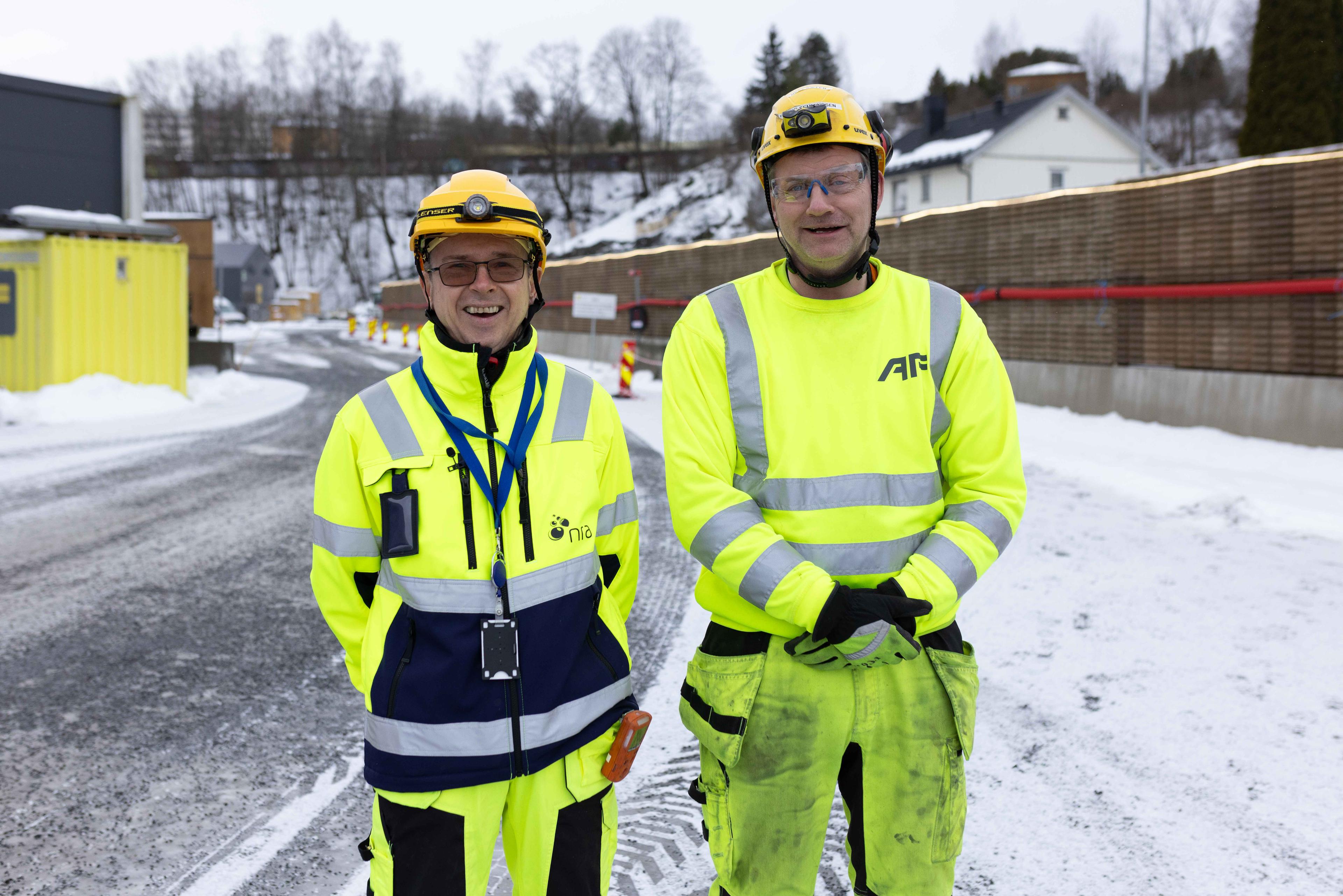 To menn i signalgule anleggsklær og hjelm står smilende ved siden av hverandre på en utendørs anleggsplass.