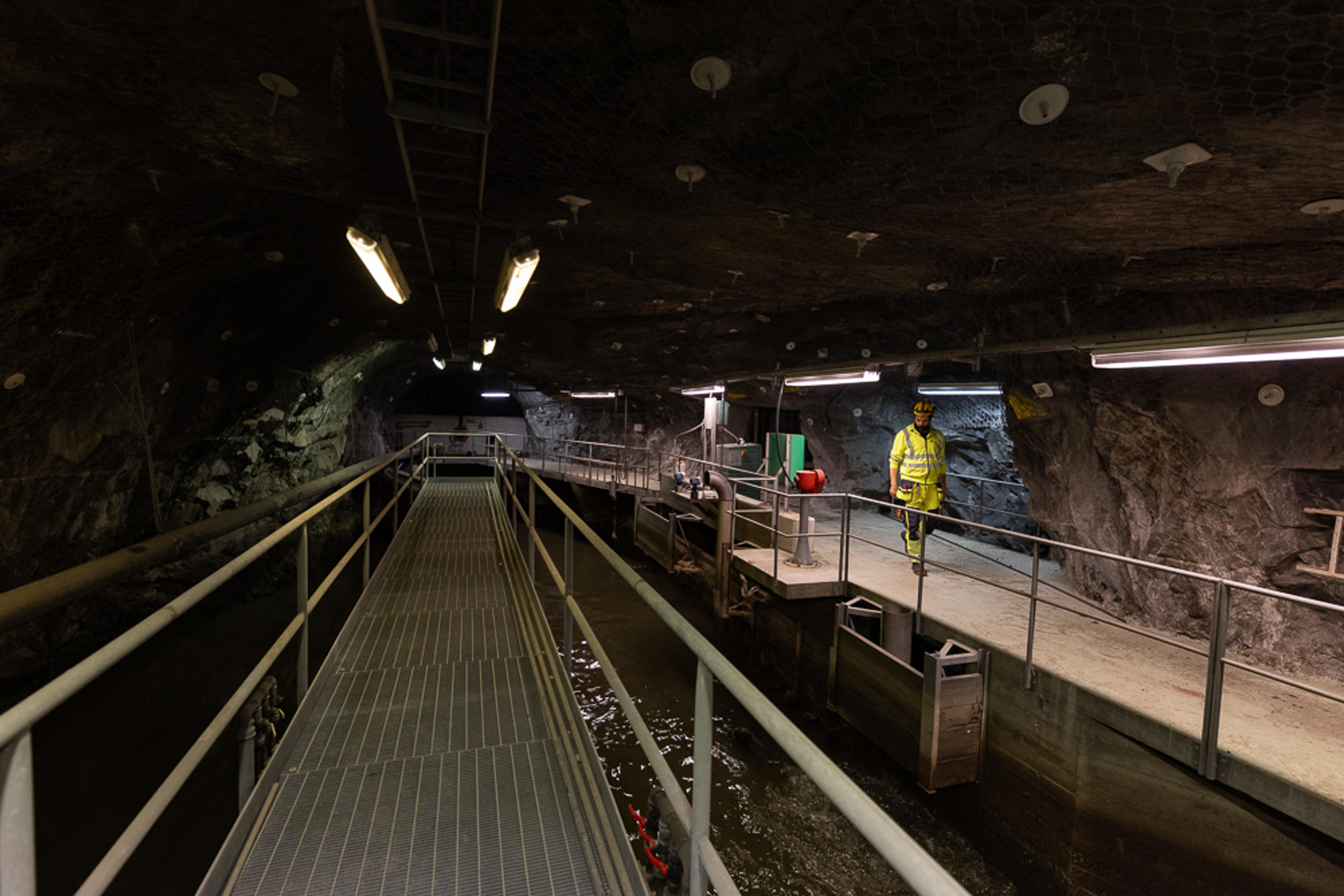 Person i gul refleksbekledning som står på en metallplattform i underjordisk tunnel med vannkanal og gangbro.