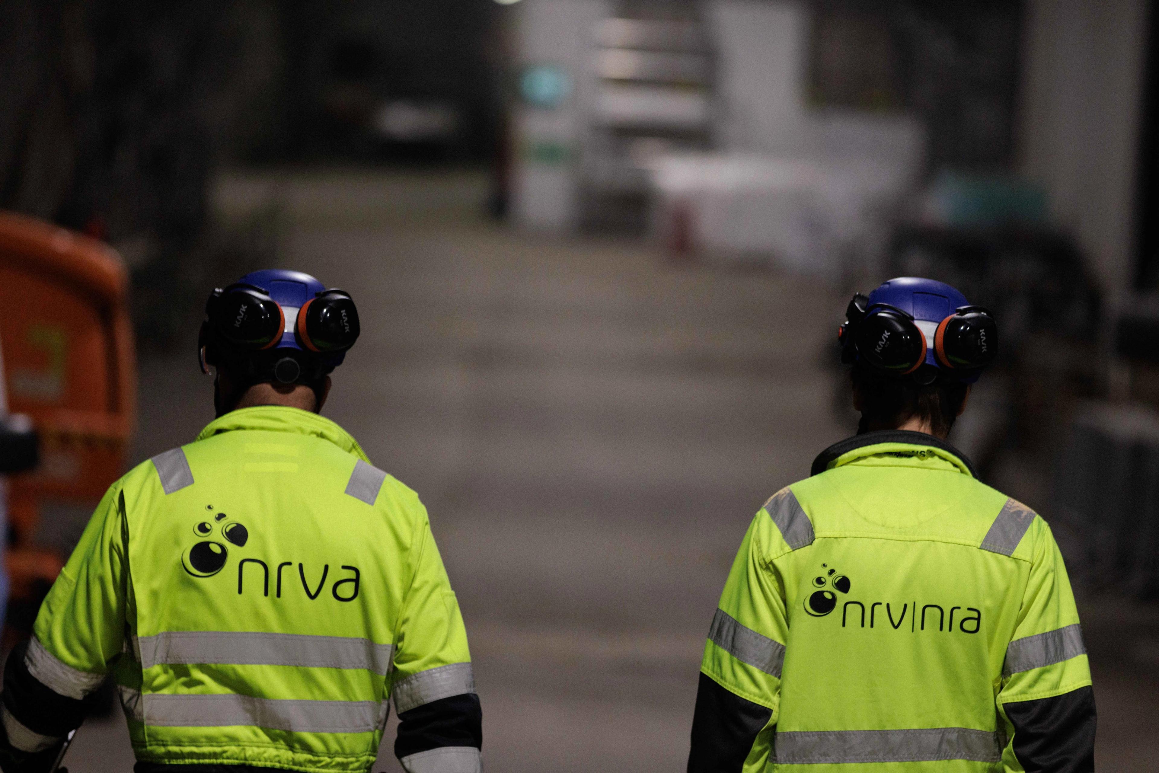 Ryggene til to mennesker med vernehjelm og signalgul jakke med NRVA-logo.