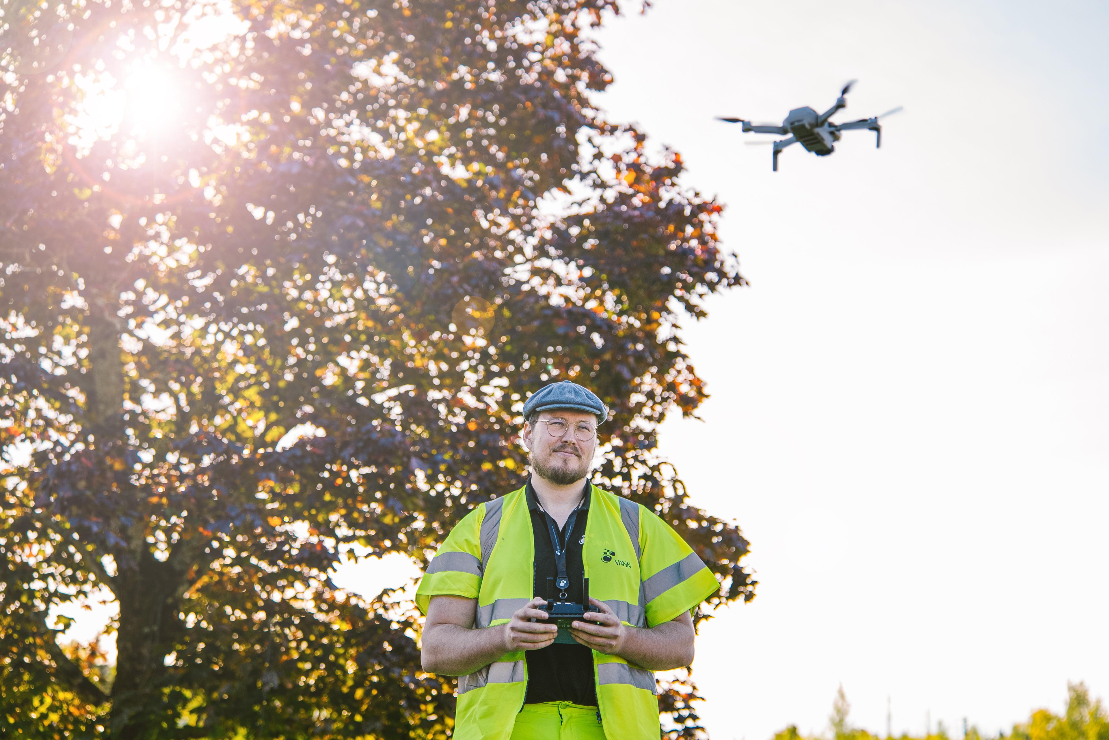 Mann i refleksvest styrer drone, høsttre i bakgrunnen med solskjær