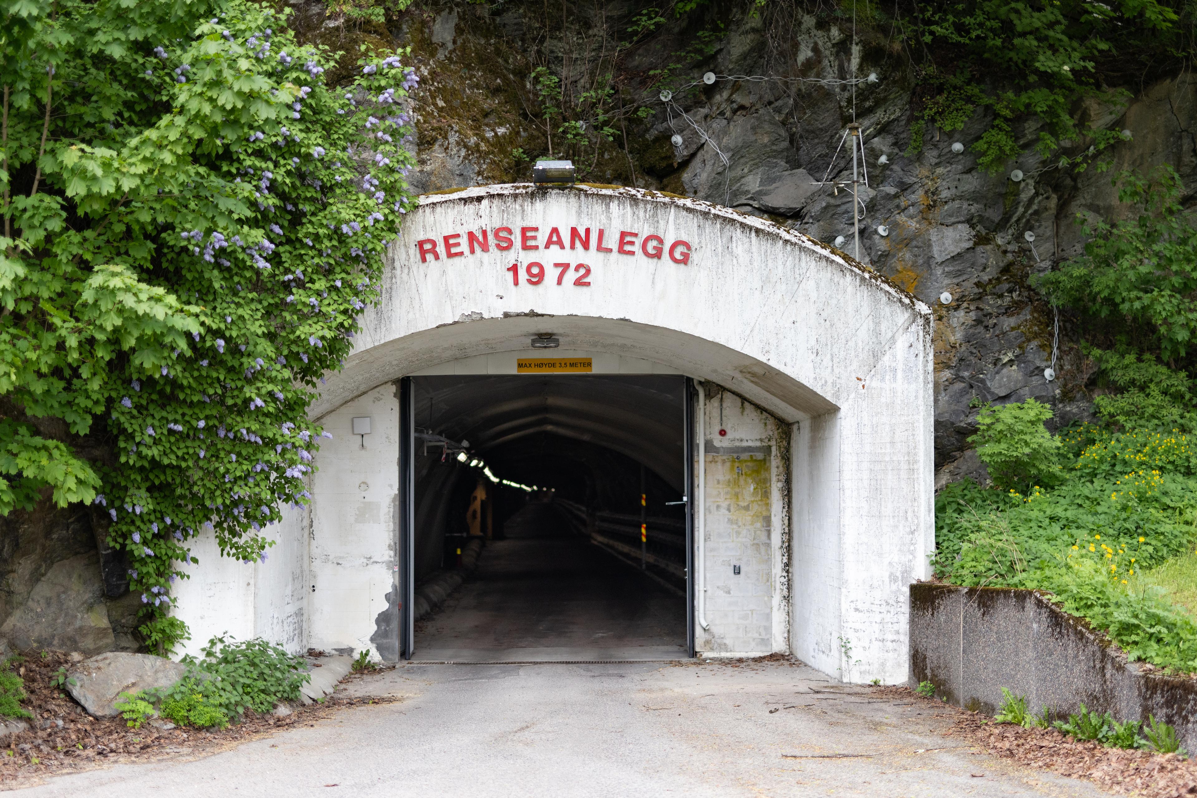 Gammel betonginngang til renseanlegg fra 1972, omkranset av grønne trær og fjell, med hvit skyvedør