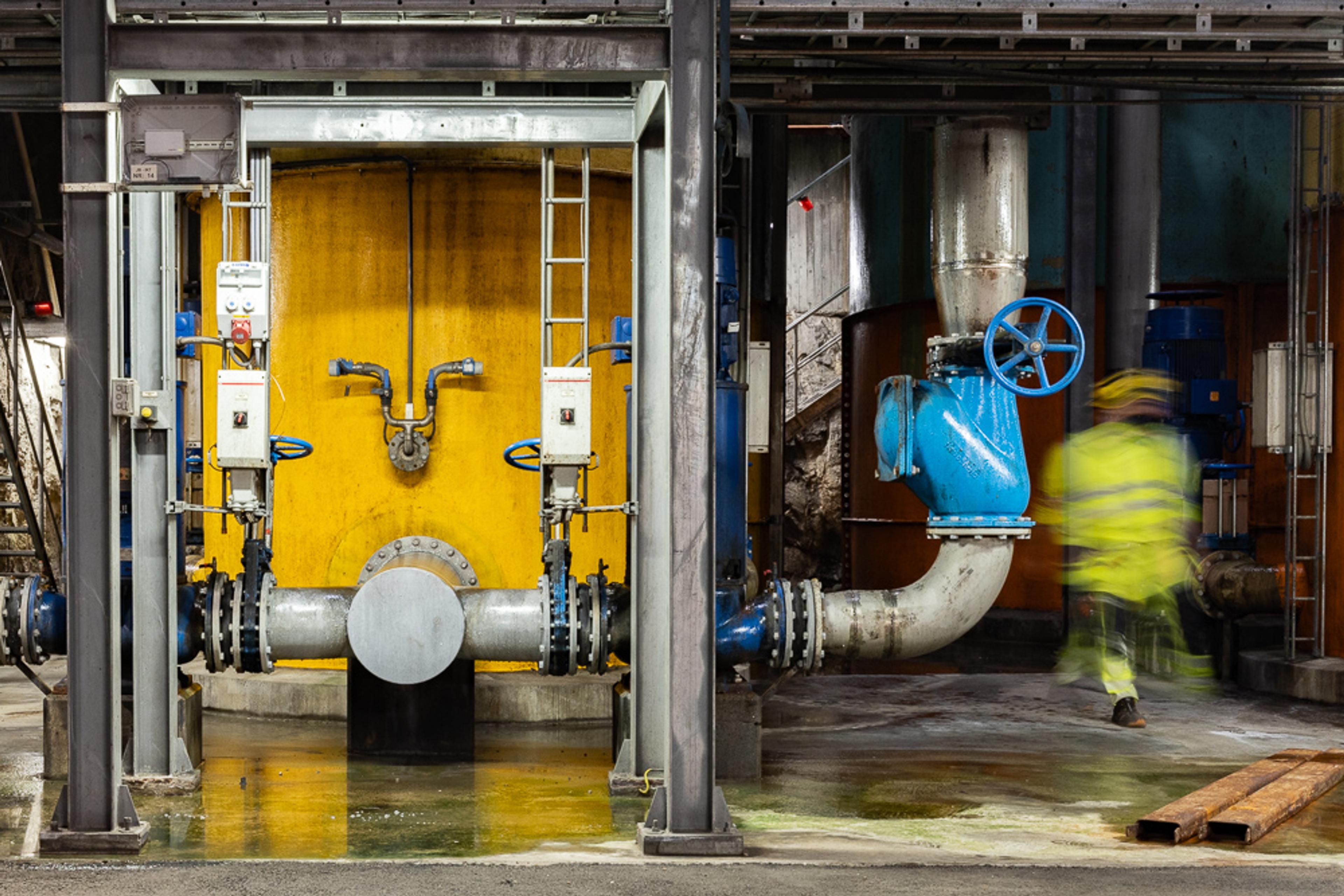 Person i gul refleksjakke i bevegelse ved vannrørsystem med blå ventil og gul vegg.