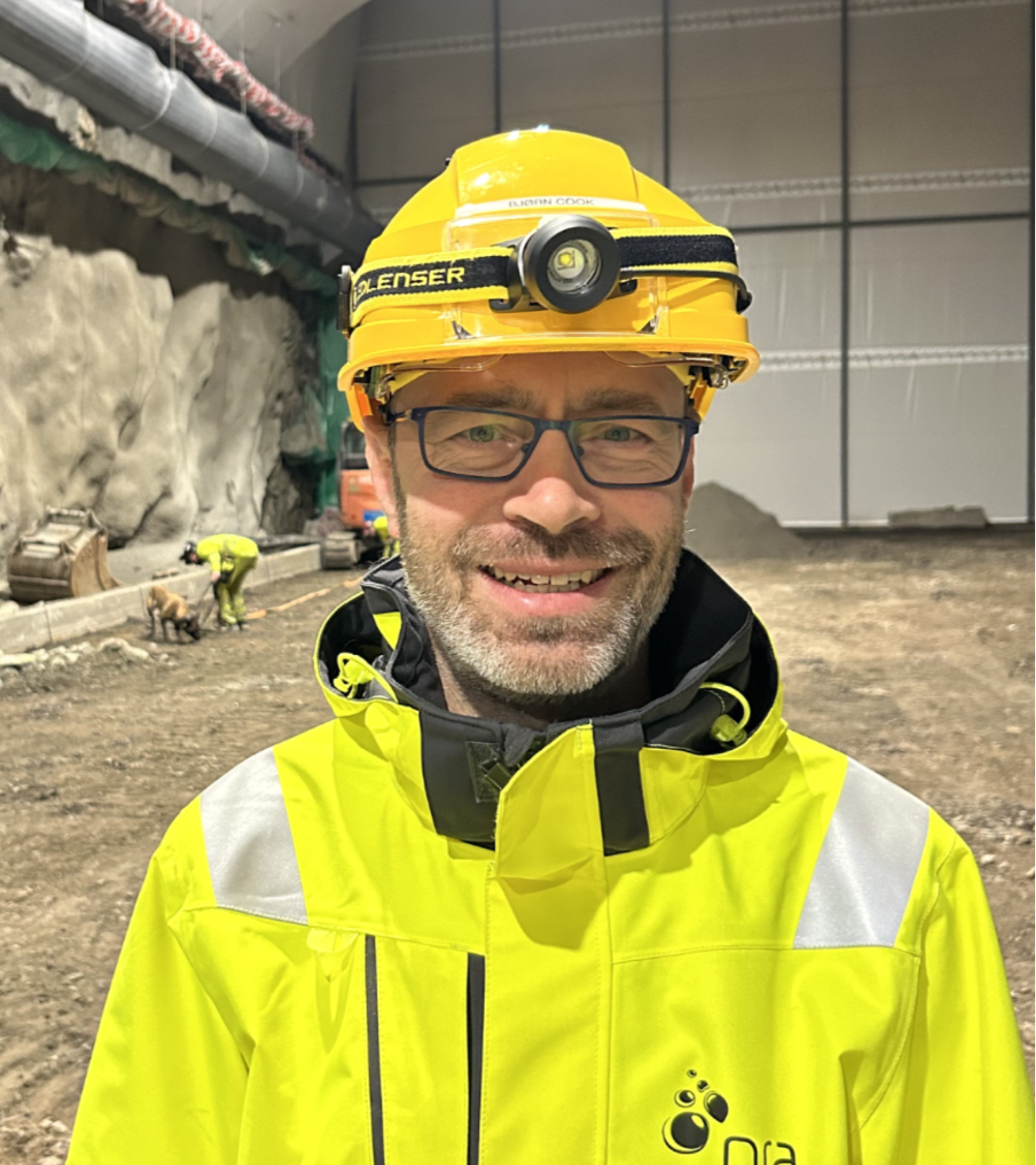 Mann med briller og skjegg som smiler mot kameraet, iført gul hjelm med hodelykt og gul refleksjakke med NRA-logo i en tunnelkonstruksjon.