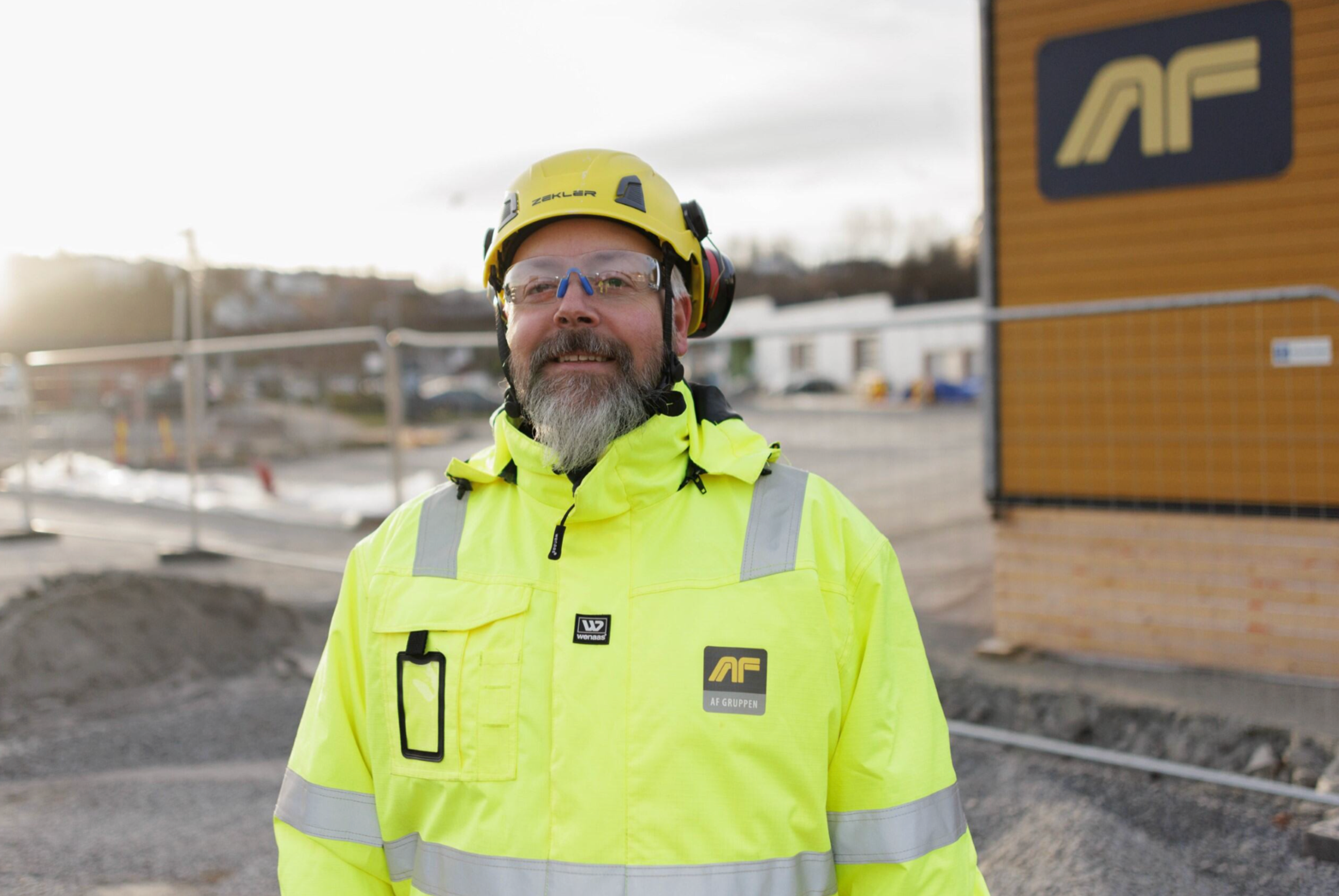 Mann med skjegg, vernebriller, hjelm og signalgul arbeidsjakke står smilende på utendørs anleggsområde.