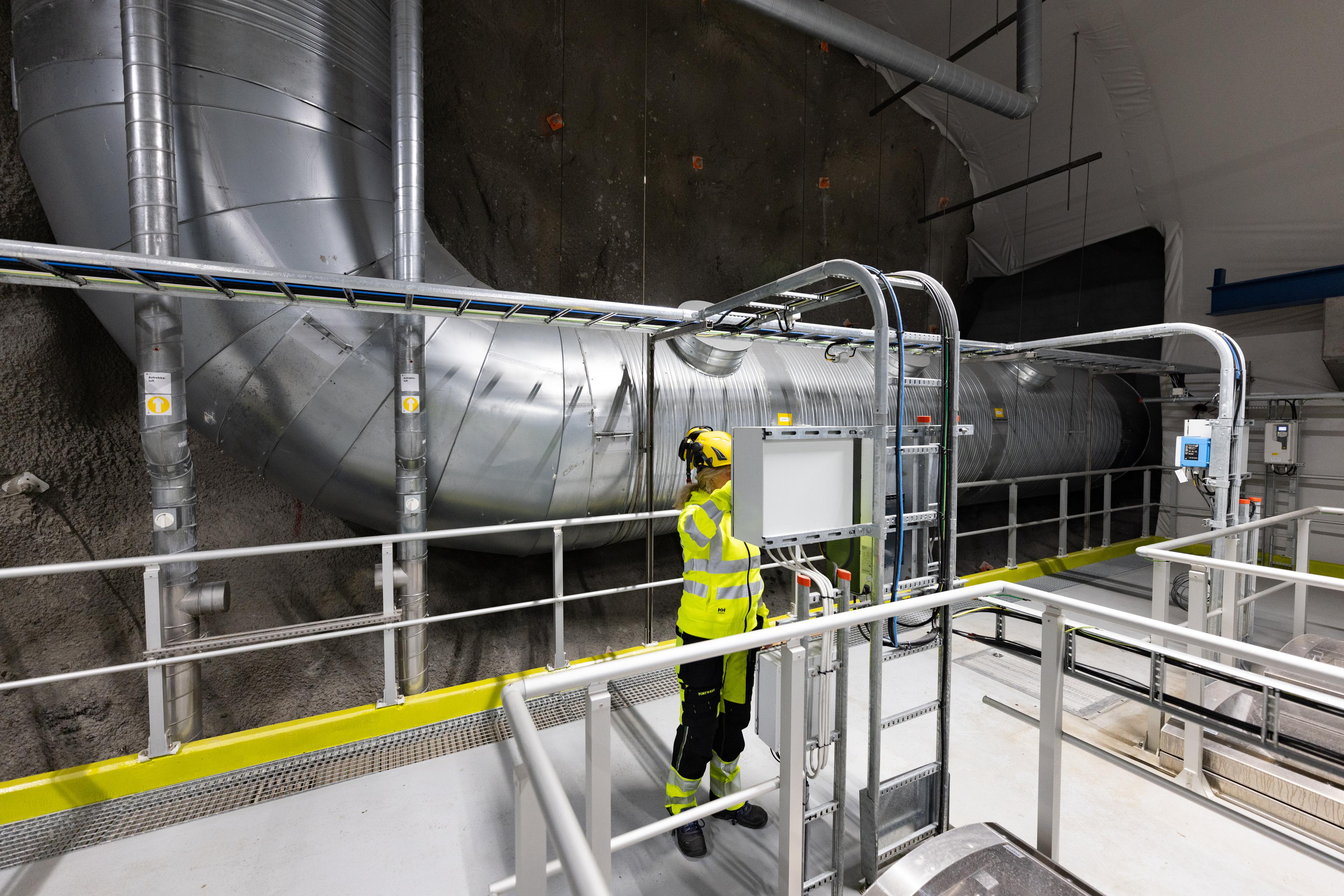 Ansatt i refleksvest inspiserer stor sølvfarget rørinstallasjon inne i industriell hall med betongvegger