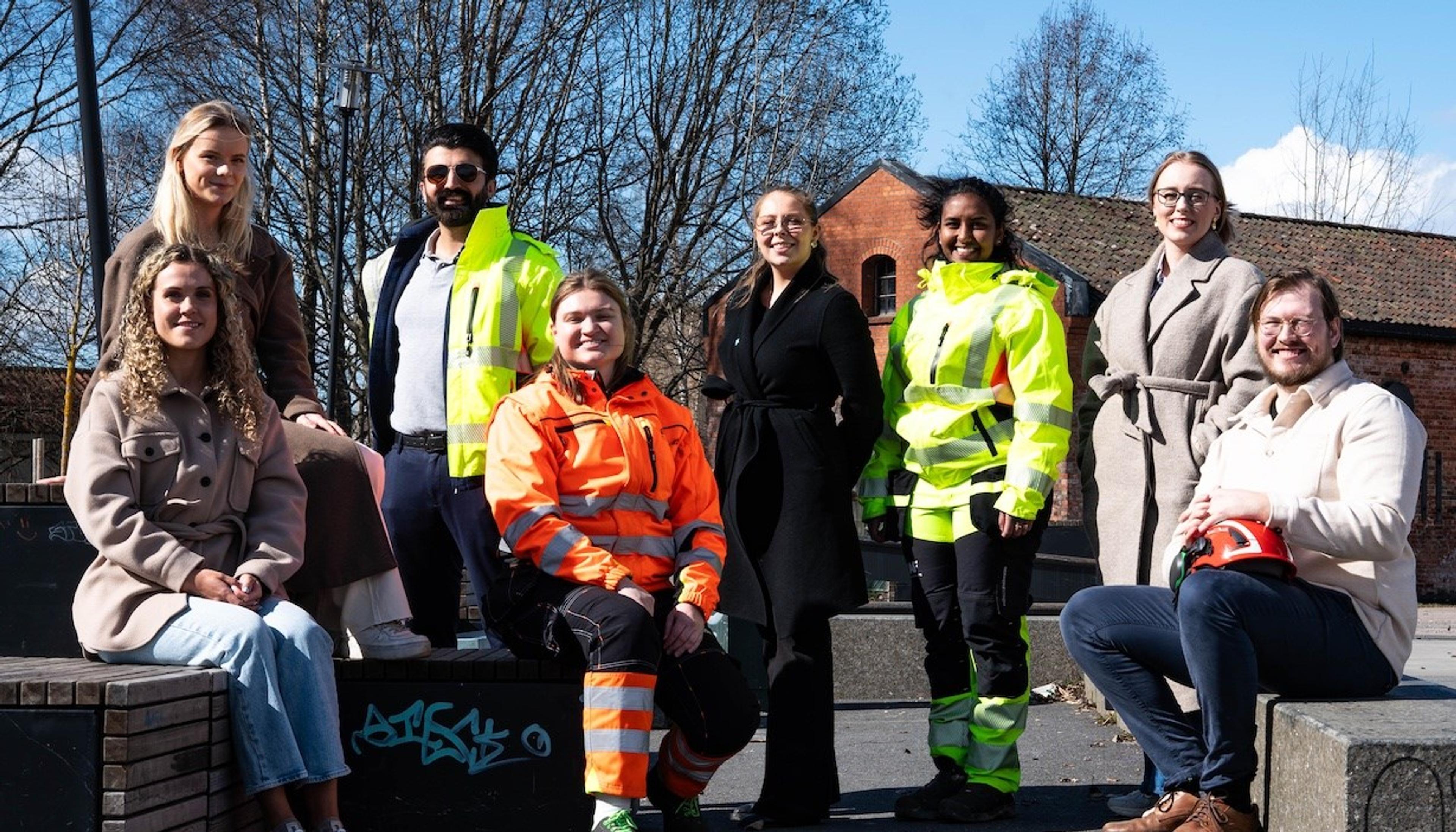 Gruppe av unge arbeidstakere eller praktikanter, noen i refleksvester og arbeidsklær, andre i vanlige klær, som poserer utendørs på en solrik dag.