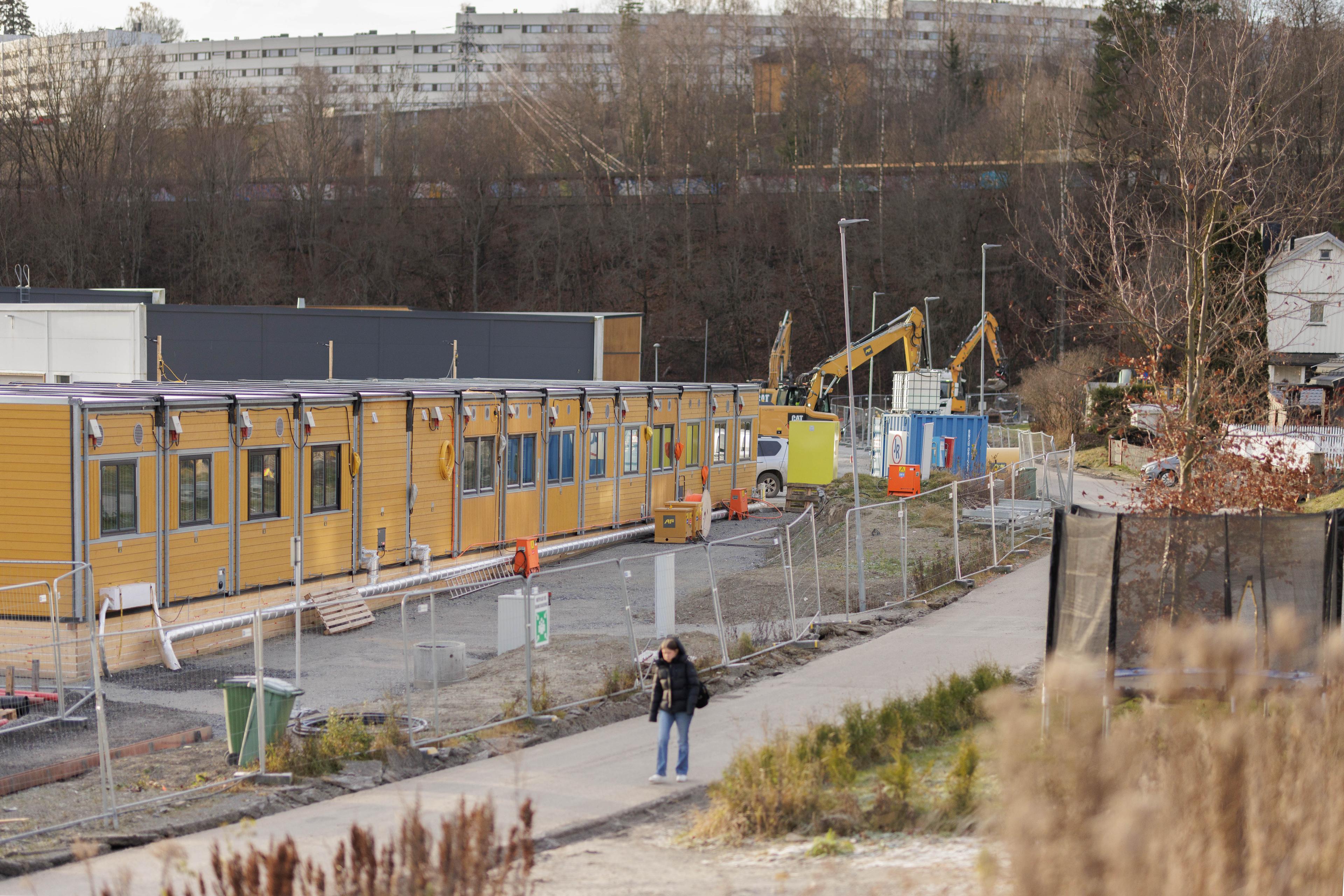 Byggeplass med gule modulbygg og anleggsmaskiner, med en person som går på gangvei og boligblokker synlige i bakgrunnen.