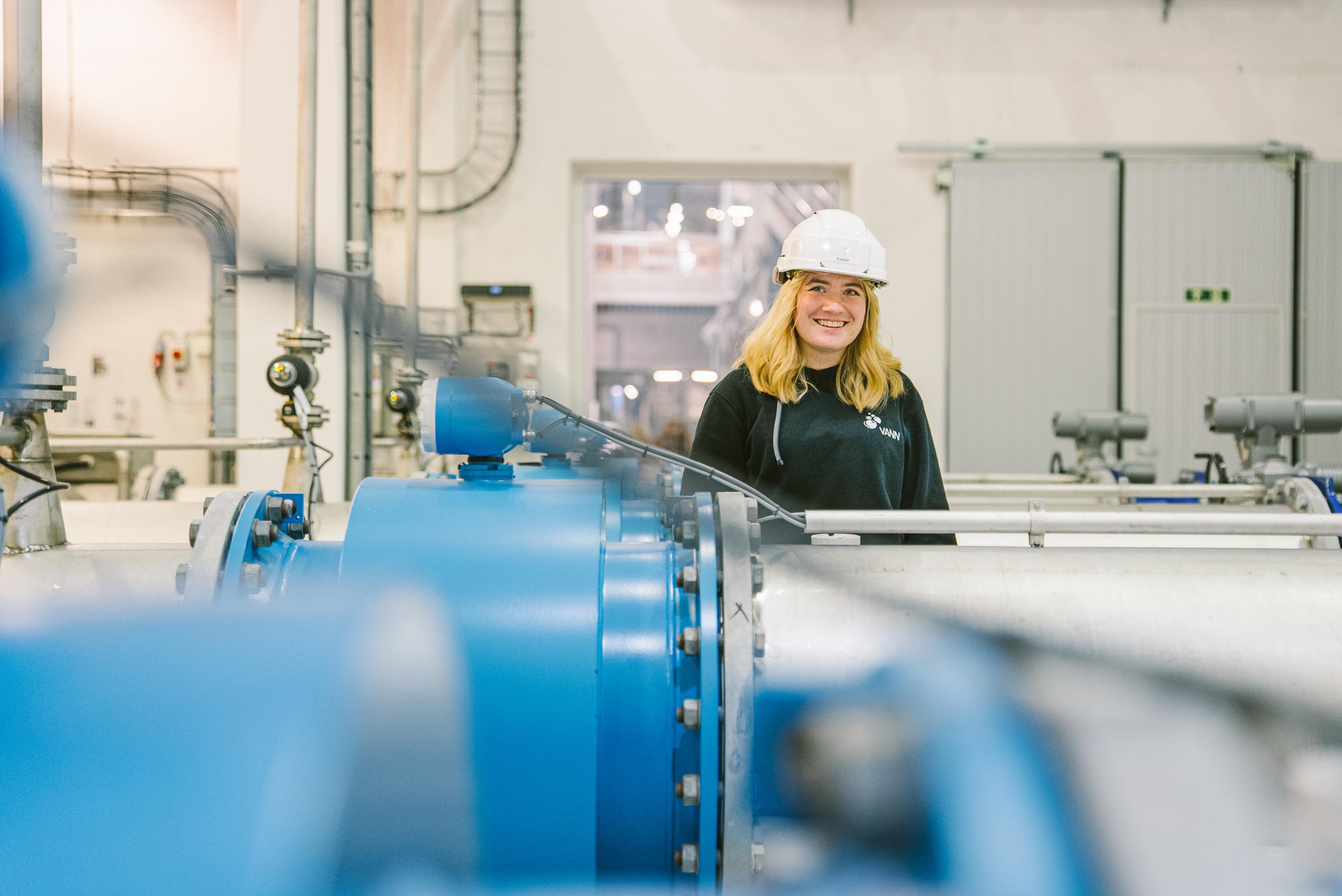 Smilende ansatt i Vann-logo-genser og hvit hjelm ved store blå industrielle rør og maskiner