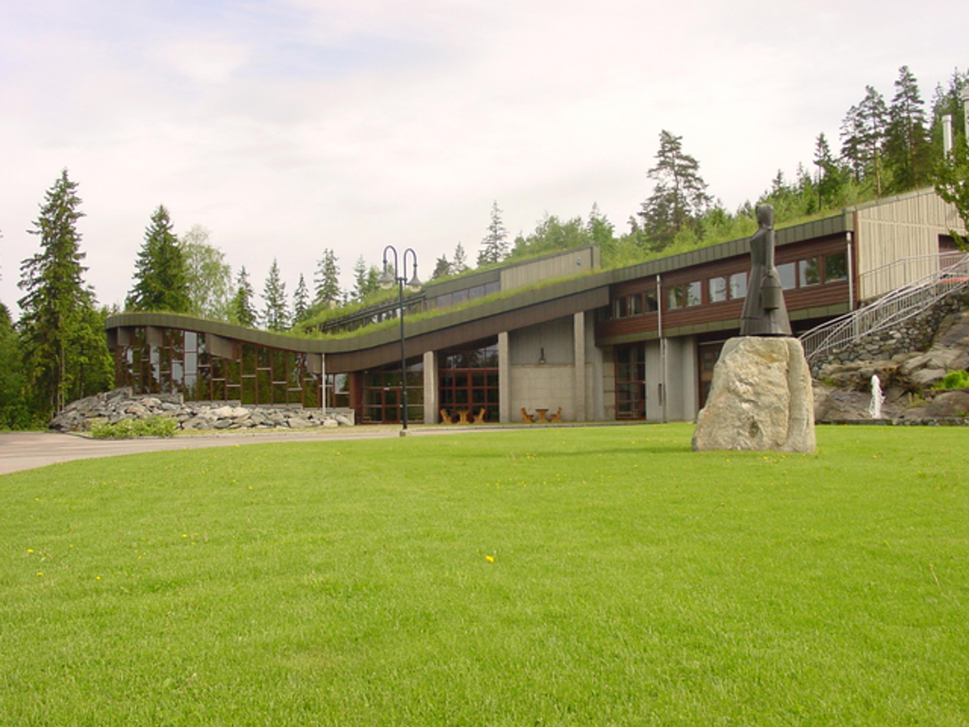 Moderne fjellhytte med gresstak og steinskulptur på grønn plen foran trær i skogen