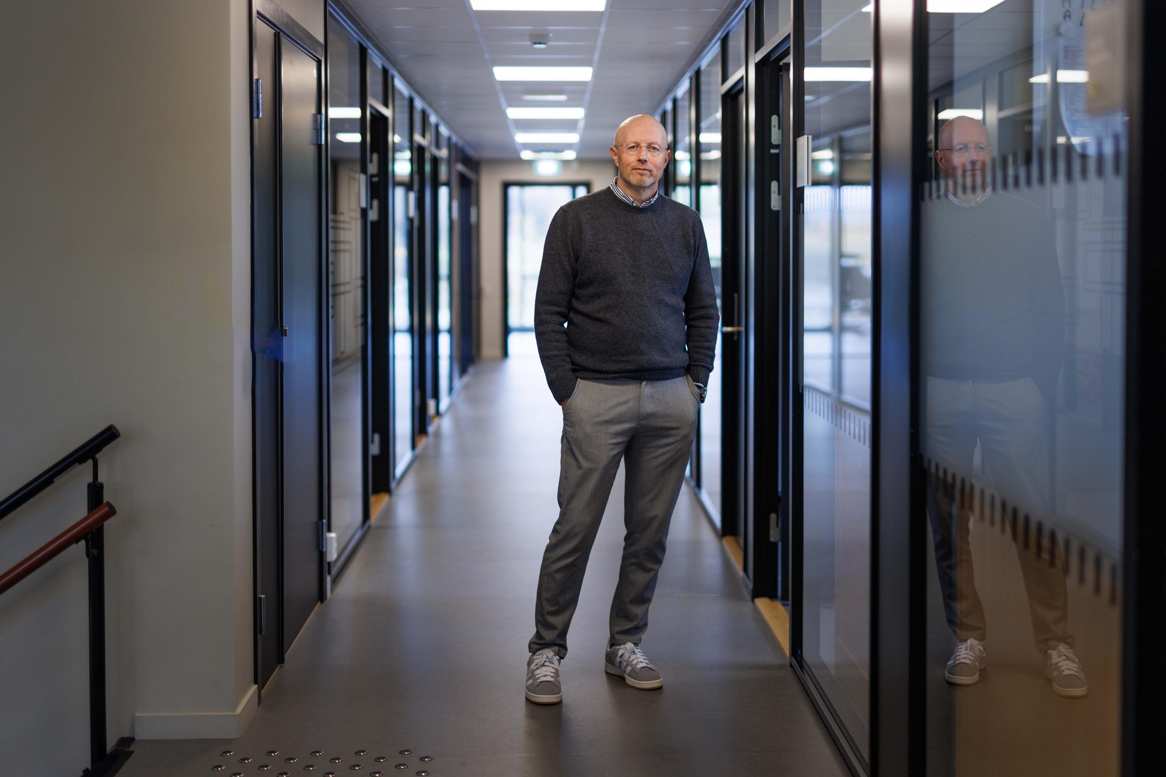 Mann i grå genser og bukser står i kontorkorridor med glassvegger