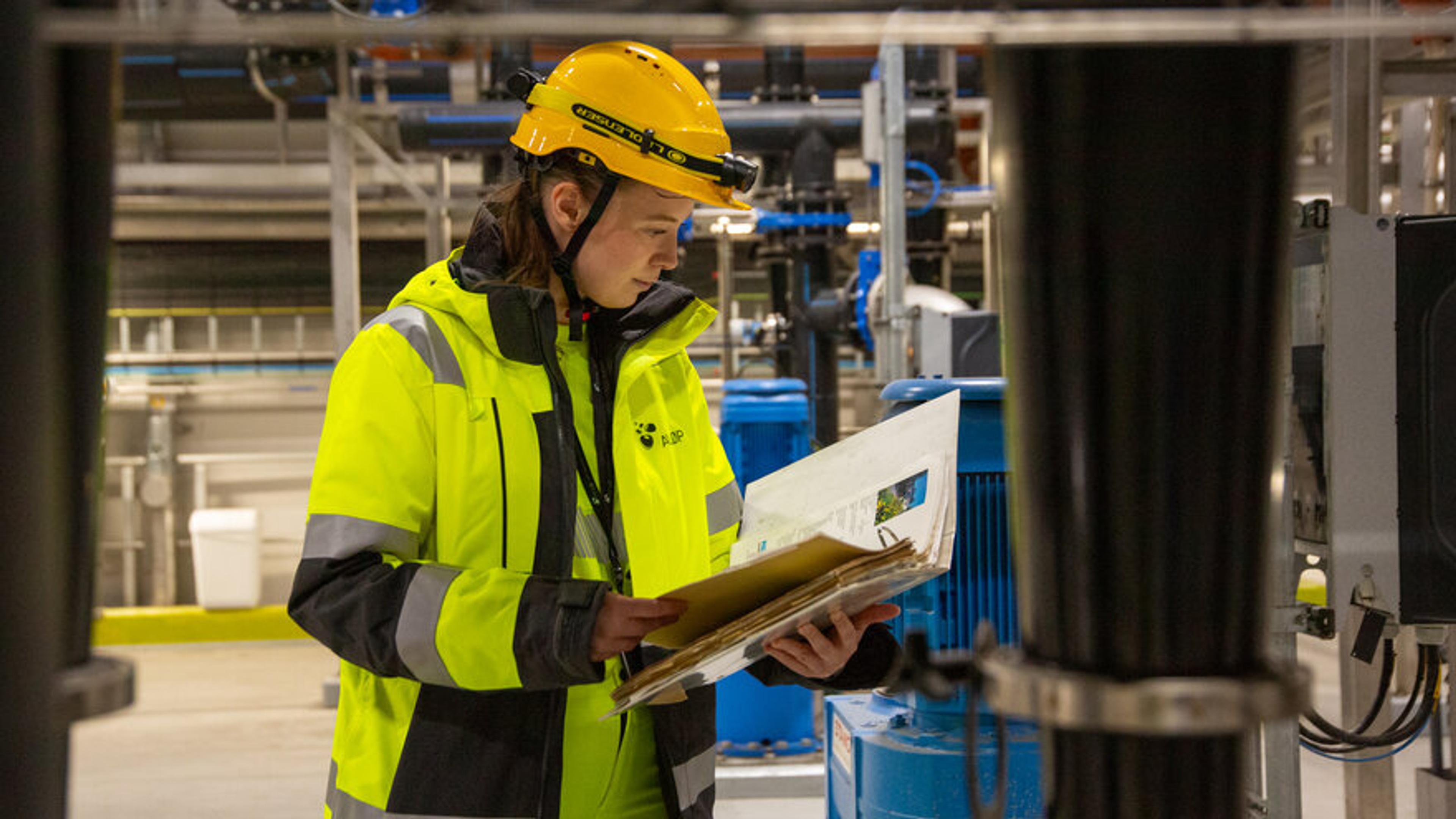 Kvinne med vernehjelm står i renseanlegg og ser ned i en perm.