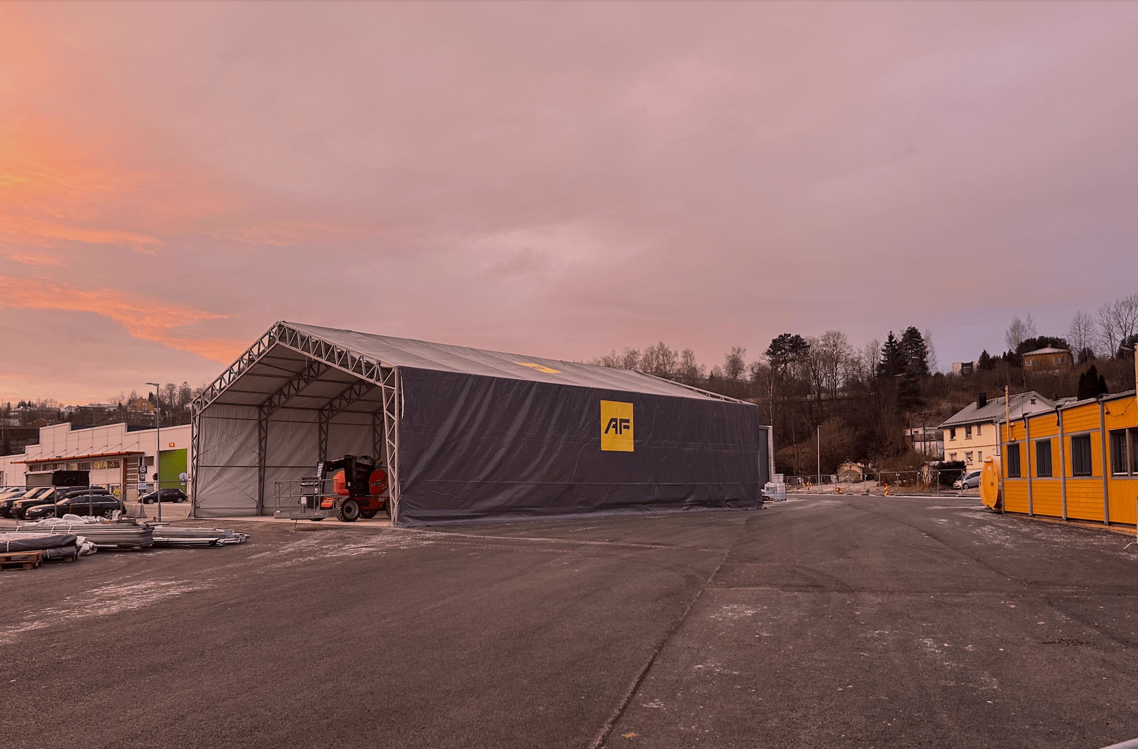 Stort, midlertidig lagertelthall med AF-logo på byggeplass i solnedgang med rosa himmel.