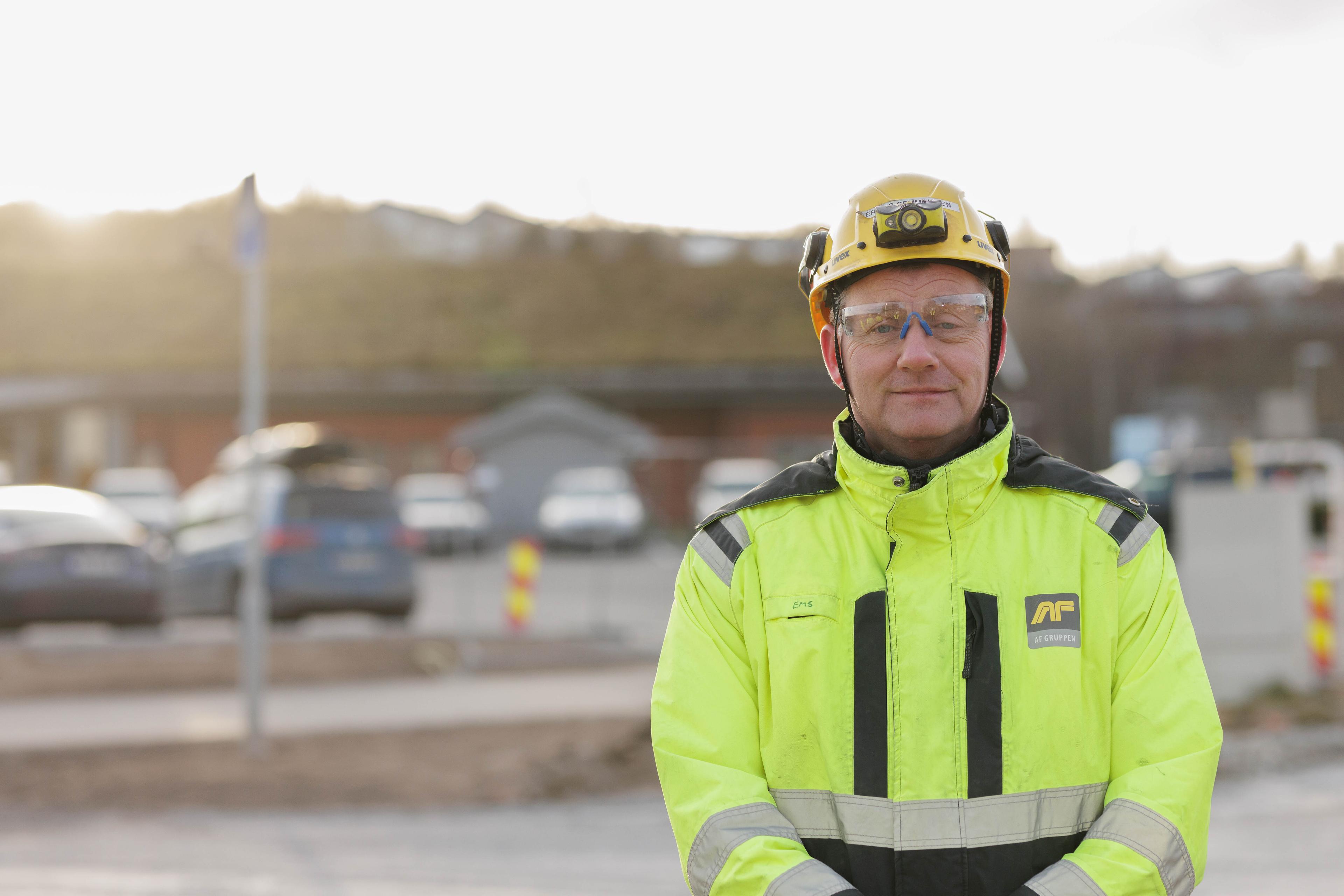 Mann med briller og gul hjelm iført fluorescerende arbeidsjakke med refleks på en byggeplass.