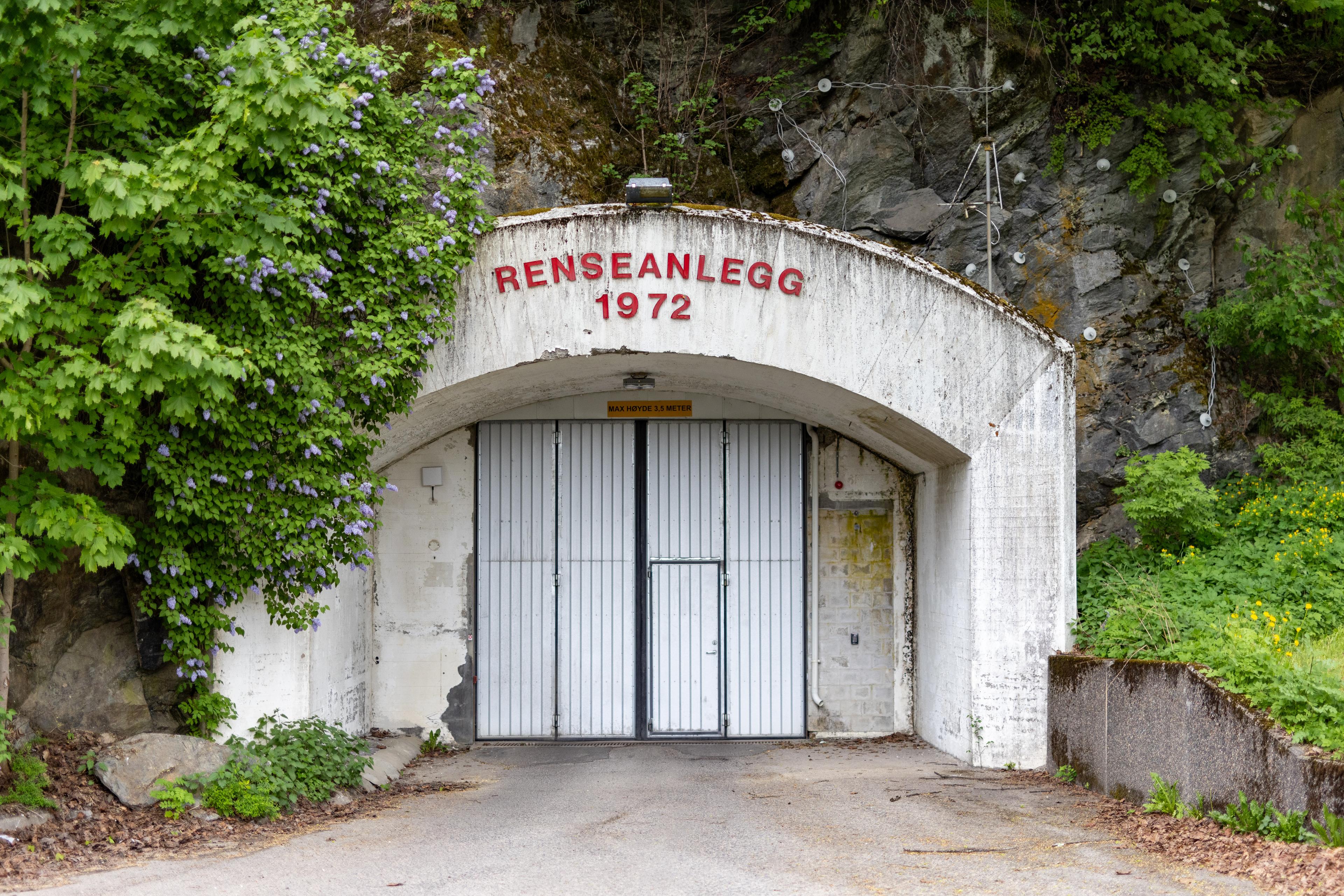 Gammel betonginngang til renseanlegg fra 1972, omkranset av grønne trær og fjell, med hvit skyvedør