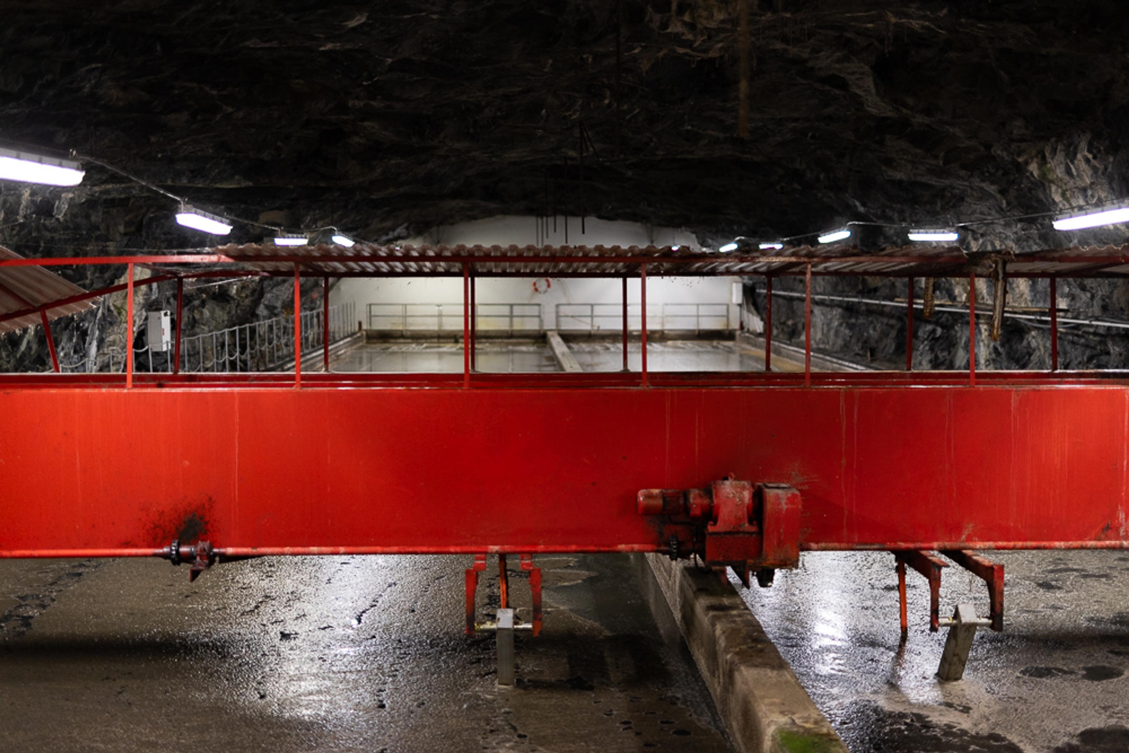 Rødt teknisk anlegg i fjellhall eller tunnelsystem med metallgelender og vannrenner i gulvet.
