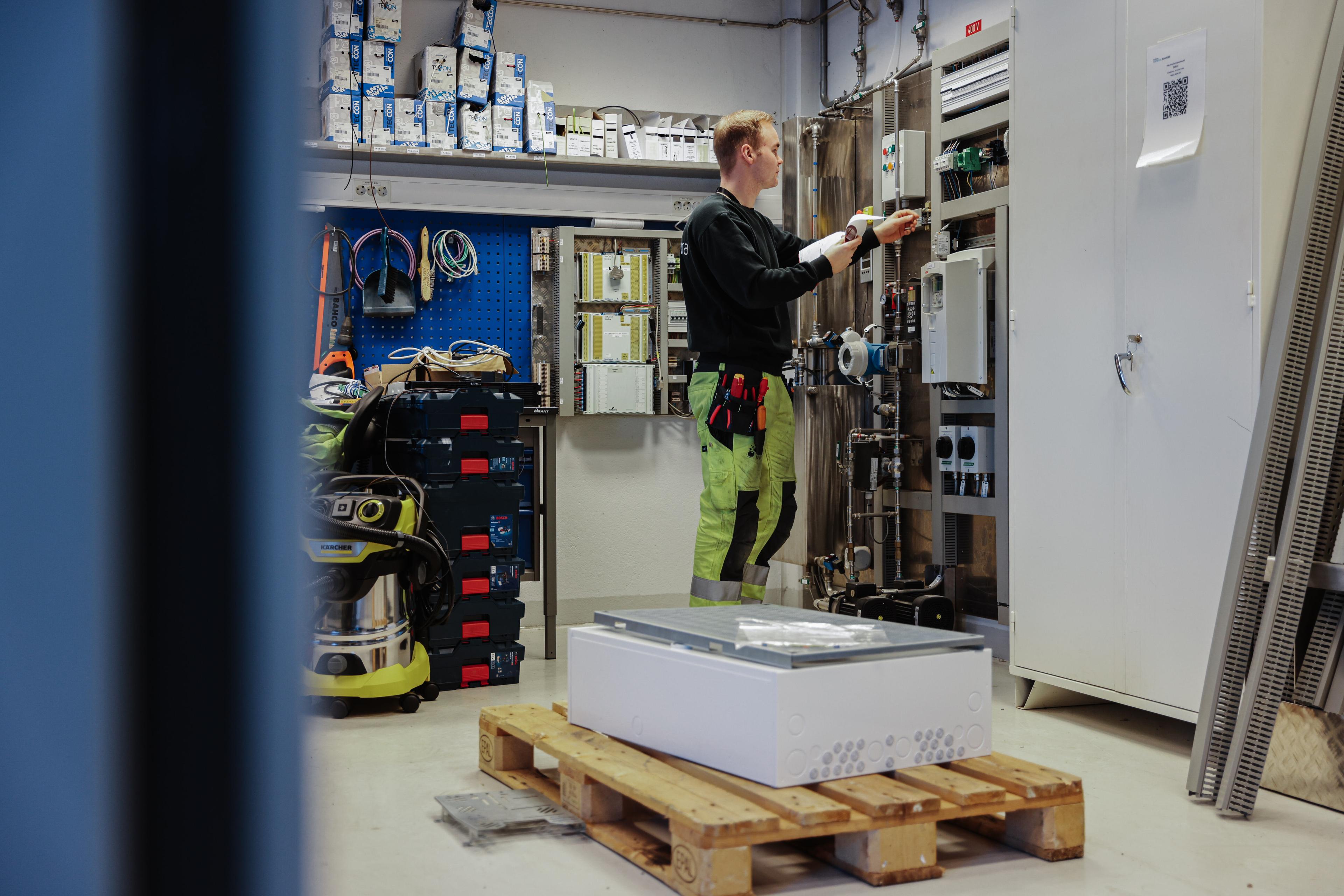 Elektriker i refleksvest jobber med elektrisk tavle i teknisk rom med verktøy, utstyr og materialer rundt seg.