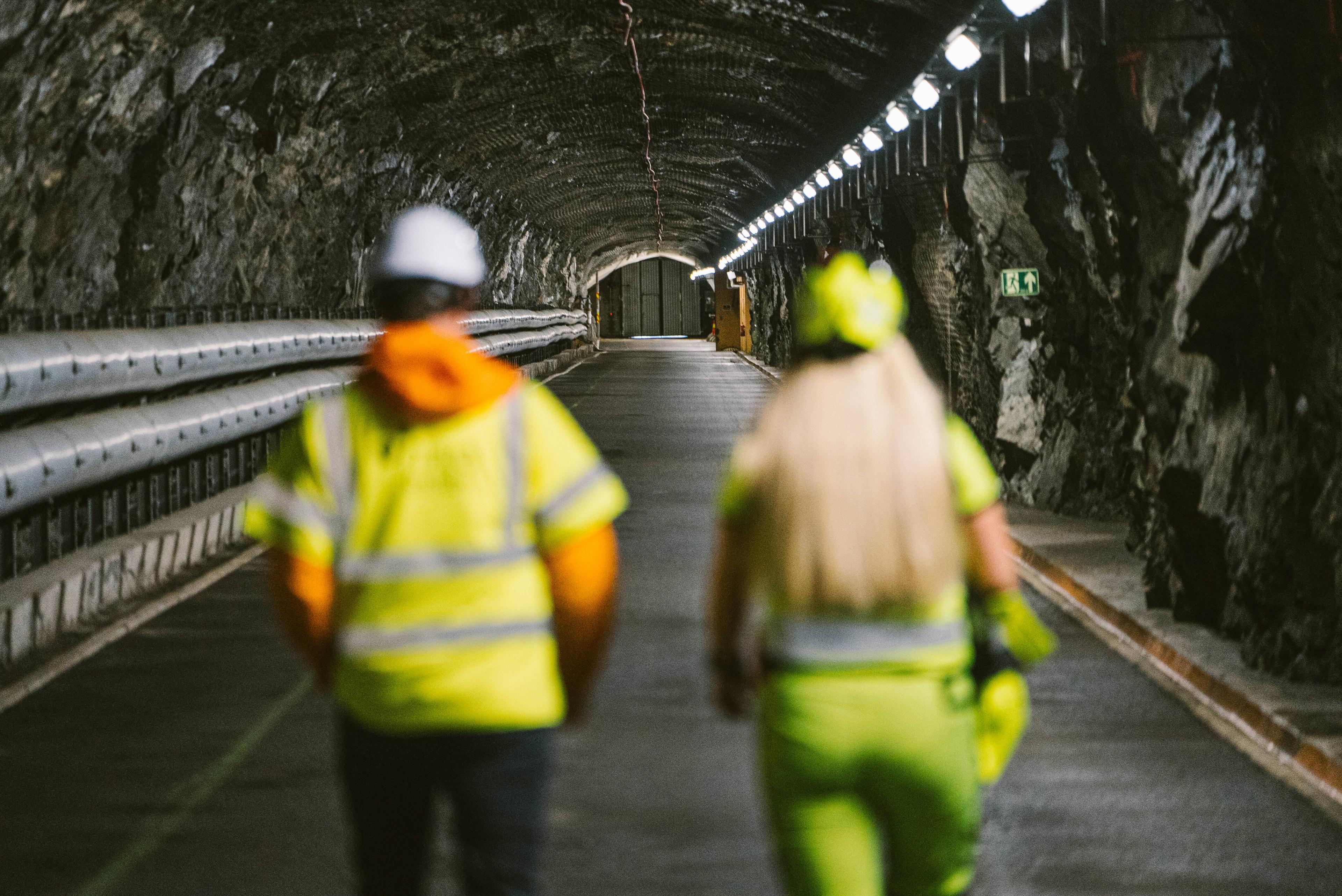To personer med refleksvester og hjelmer som går gjennom en opplyst bergtunnel med nødutgangsskilt synlig i bakgrunnen.