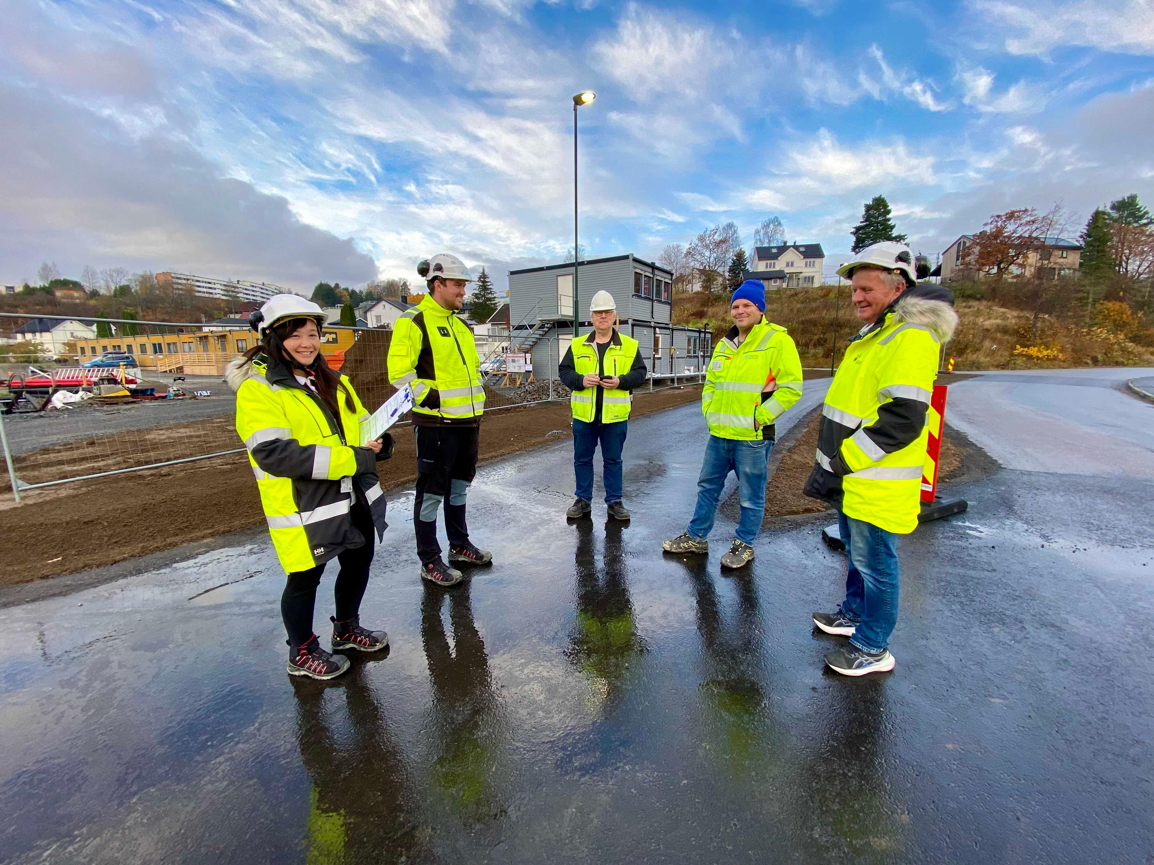 fem anleggsarbeidere i gule refleksvester og hjelmer som står på en våt asfaltert vei på et byggområde under en blå himmel med skyer.