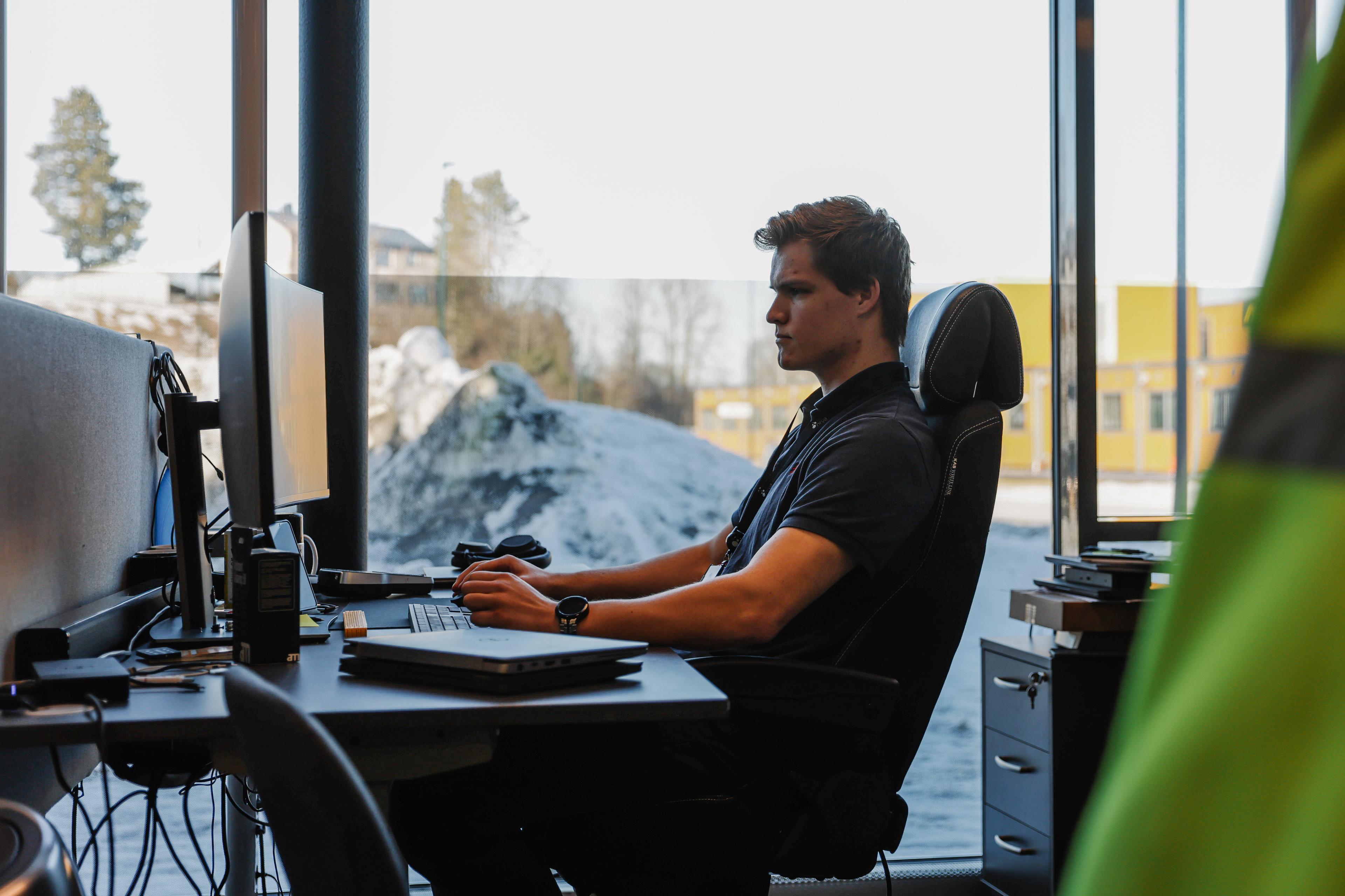 Ung mann i sort polo som arbeider ved dataskjerm i et kontor med store vinduer som viser snø utenfor.