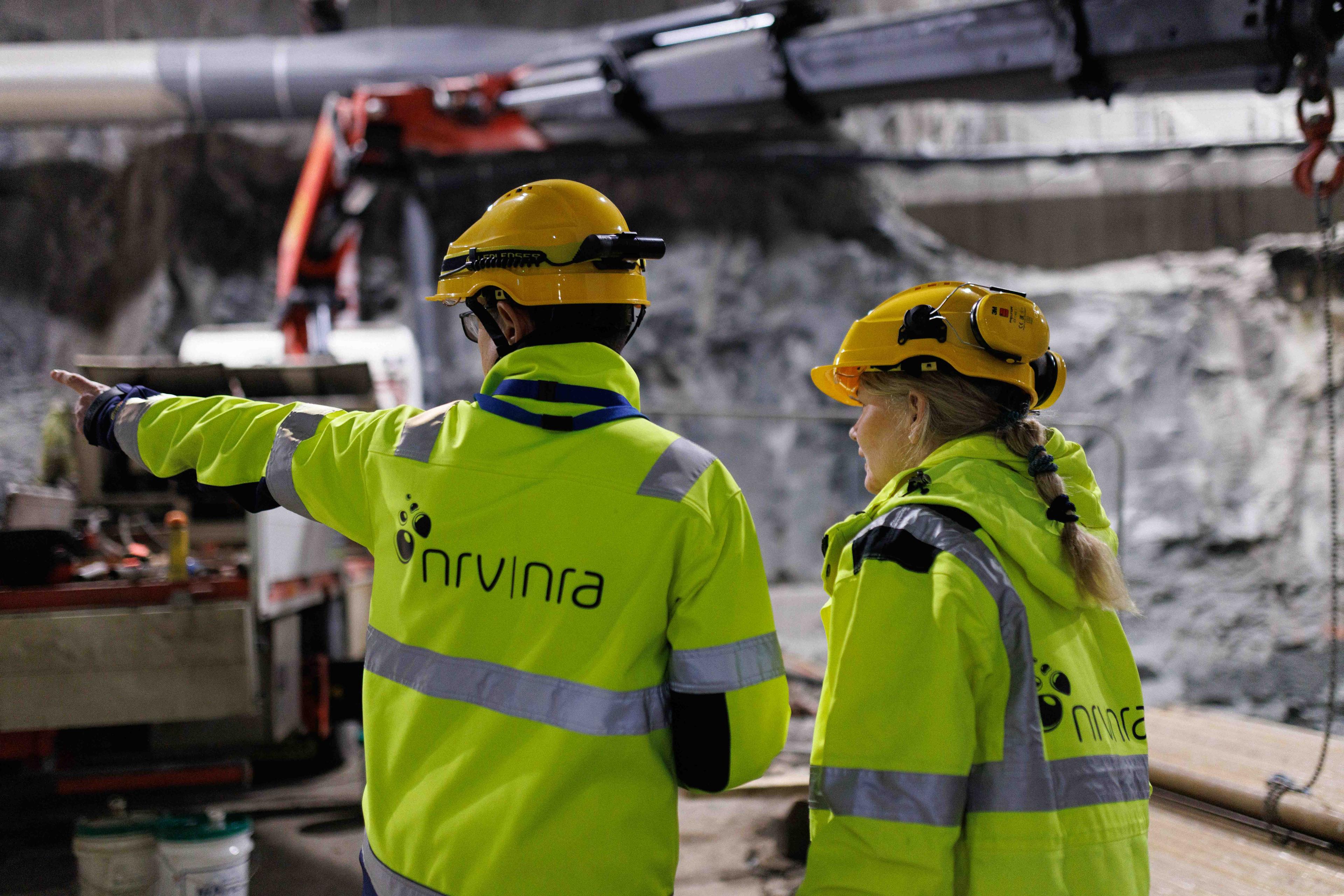 En mann og kvinne i signalgule anleggsklær og hjelm står med ryggen til inne i en tunnel og peker.