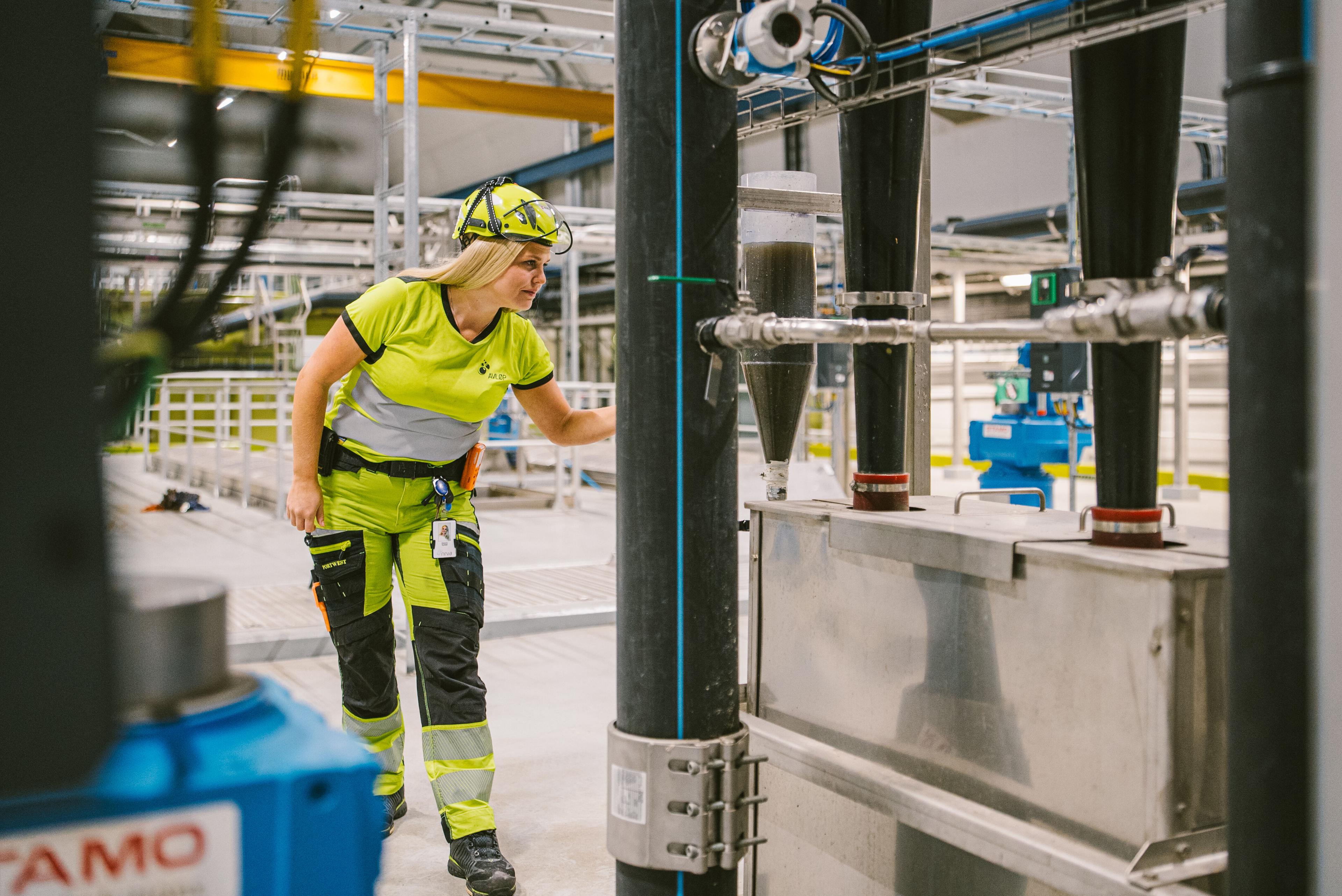 Kvinnelig arbeider med hjelm og gul arbeidsuniform som inspiserer prosessutstyr i et vannrenseanlegg.