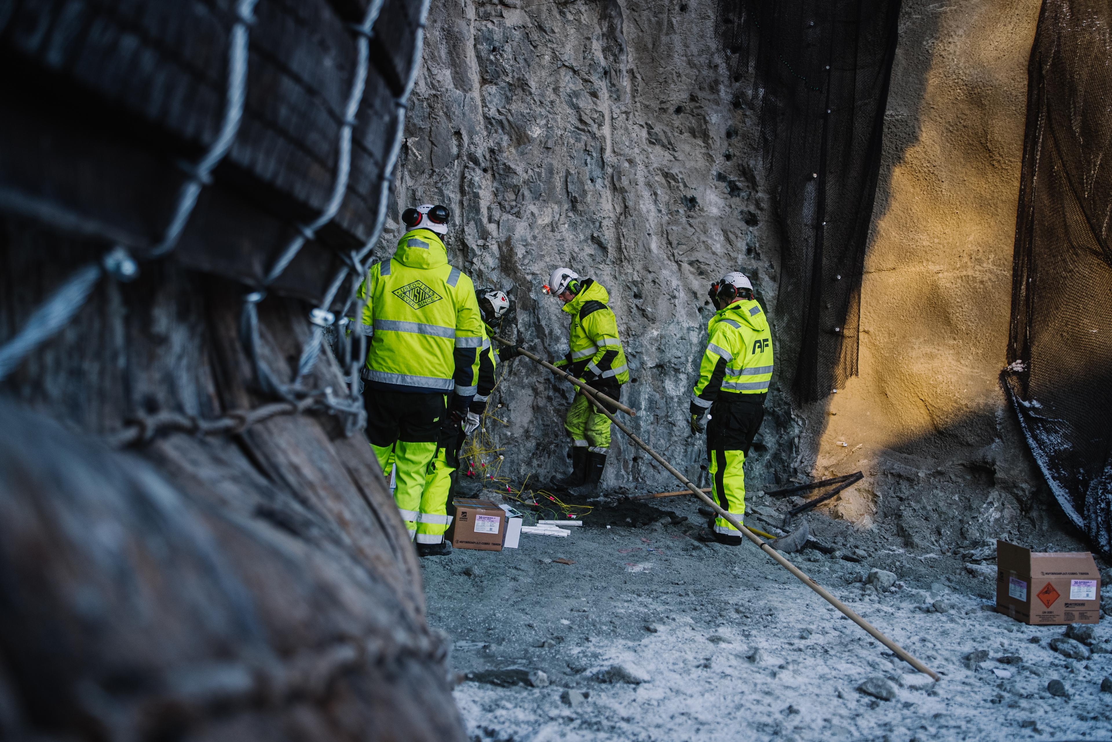 Anleggsarbeidere i gul refleksbekledning og hjelmer som arbeider ved fjellvegg i en tunnelkonstruksjon.