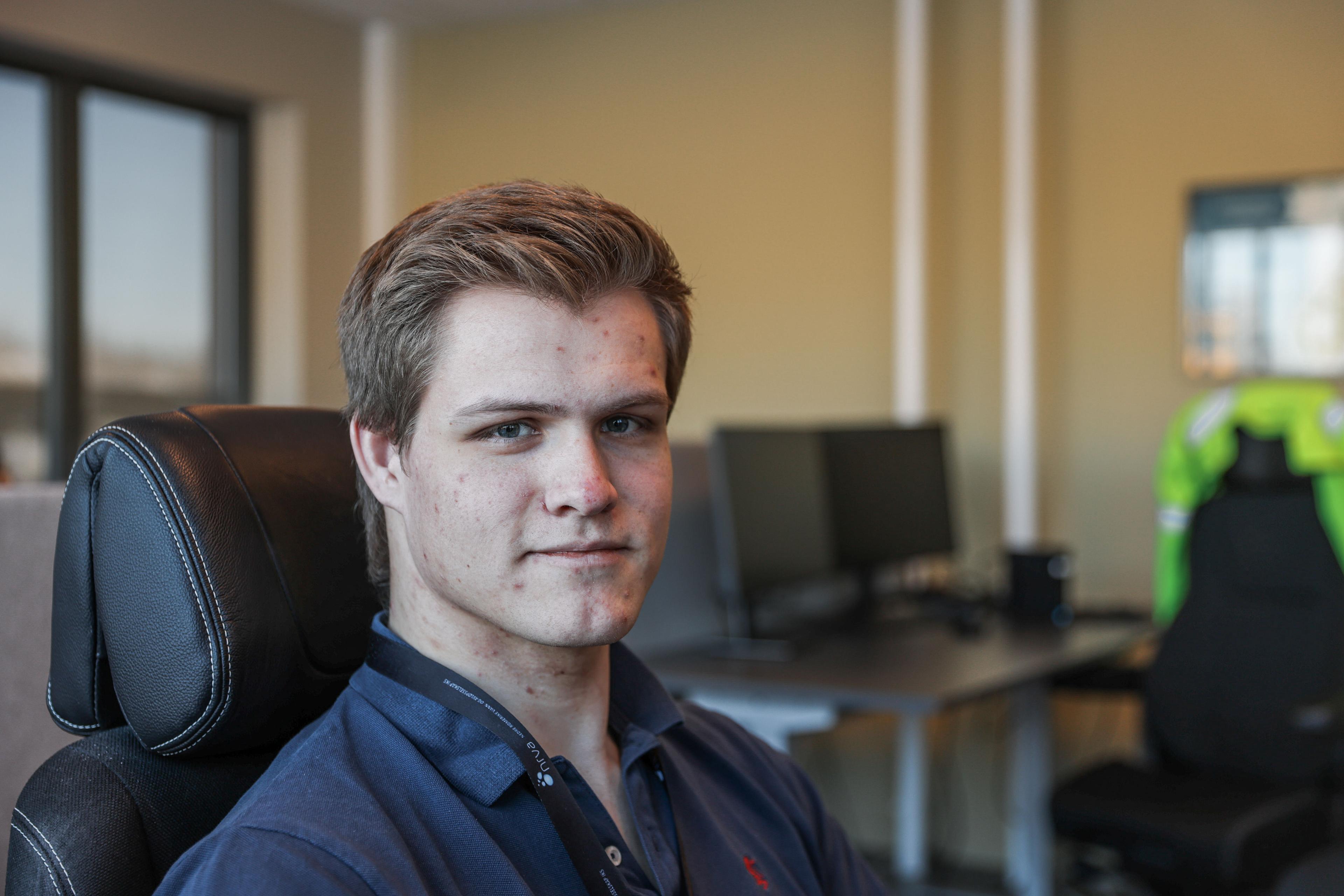 Ung mann i mørkeblå poloskjorte som sitter i kontorstol med kontorlandskap og dataskjermer i bakgrunnen.