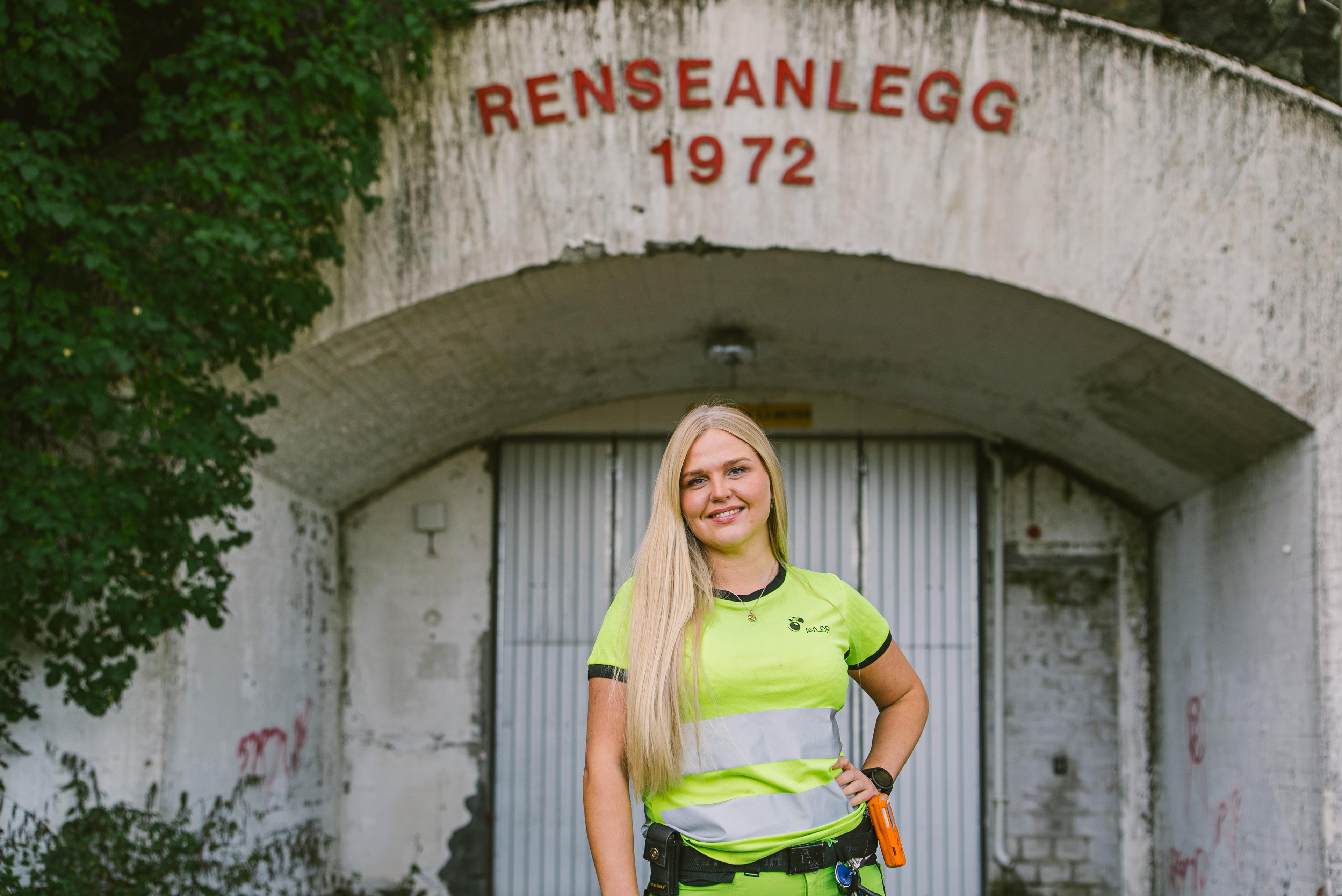 Smilende ansatt i refleksvest foran gammel renseanlegg-bygning fra 1972, med grønne trær rundt