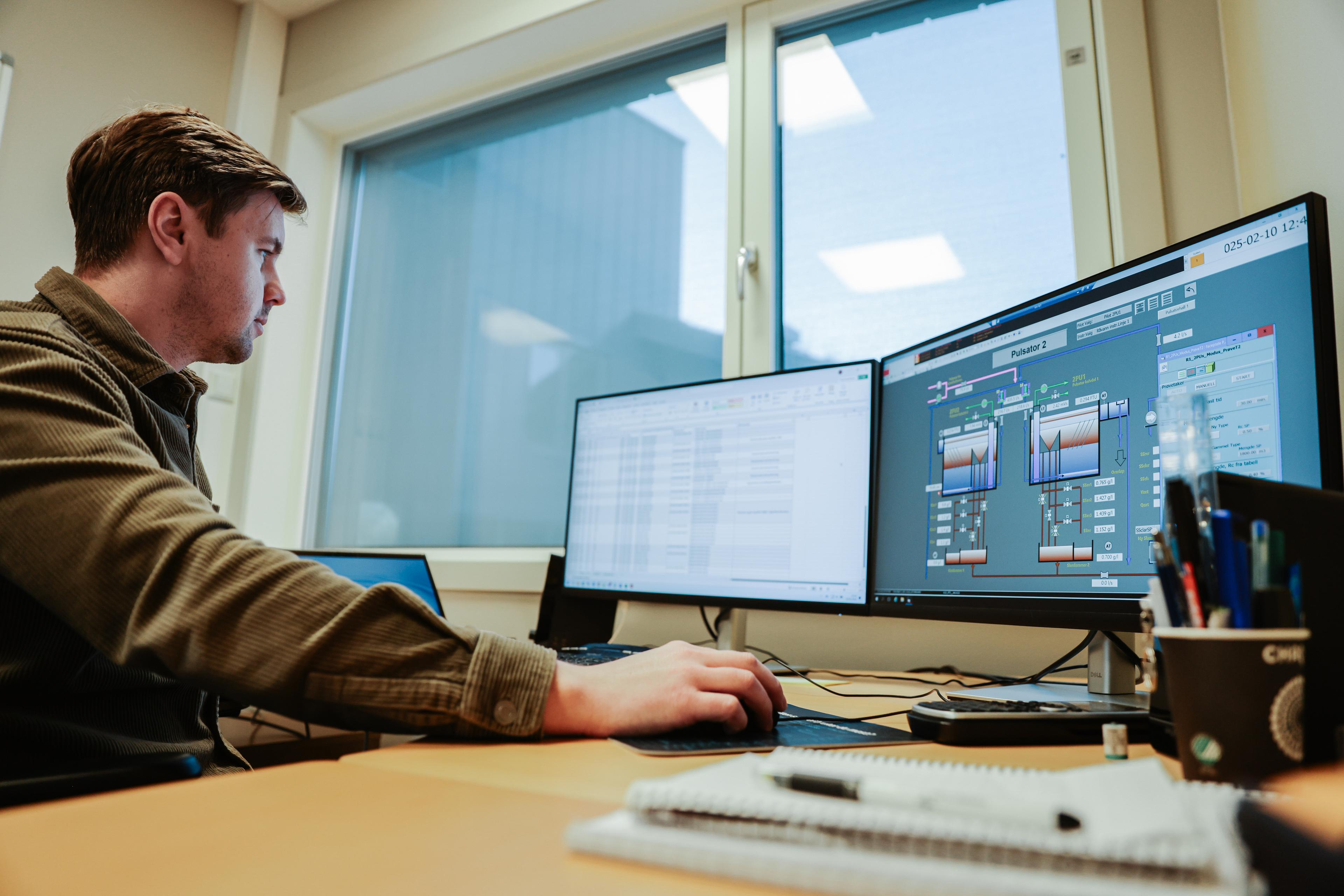 Mann i brun genser som arbeider ved skrivebord med to dataskjermer som viser tekniske data og grafer, med dagslys som strømmer inn gjennom vinduet.