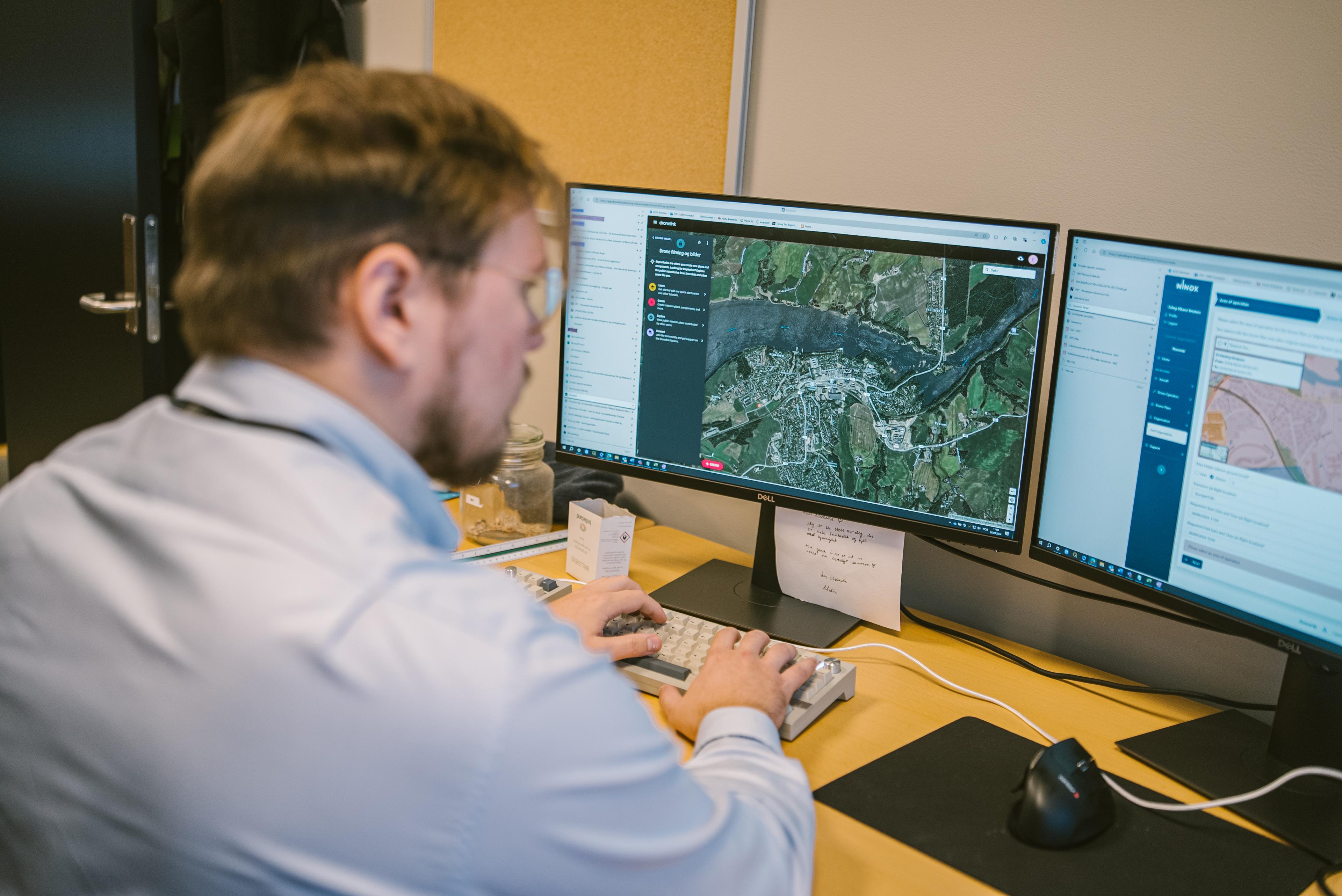 Mann i lyseblå skjorte som arbeider ved datamaskiner med kartsystemer på to skjermer.