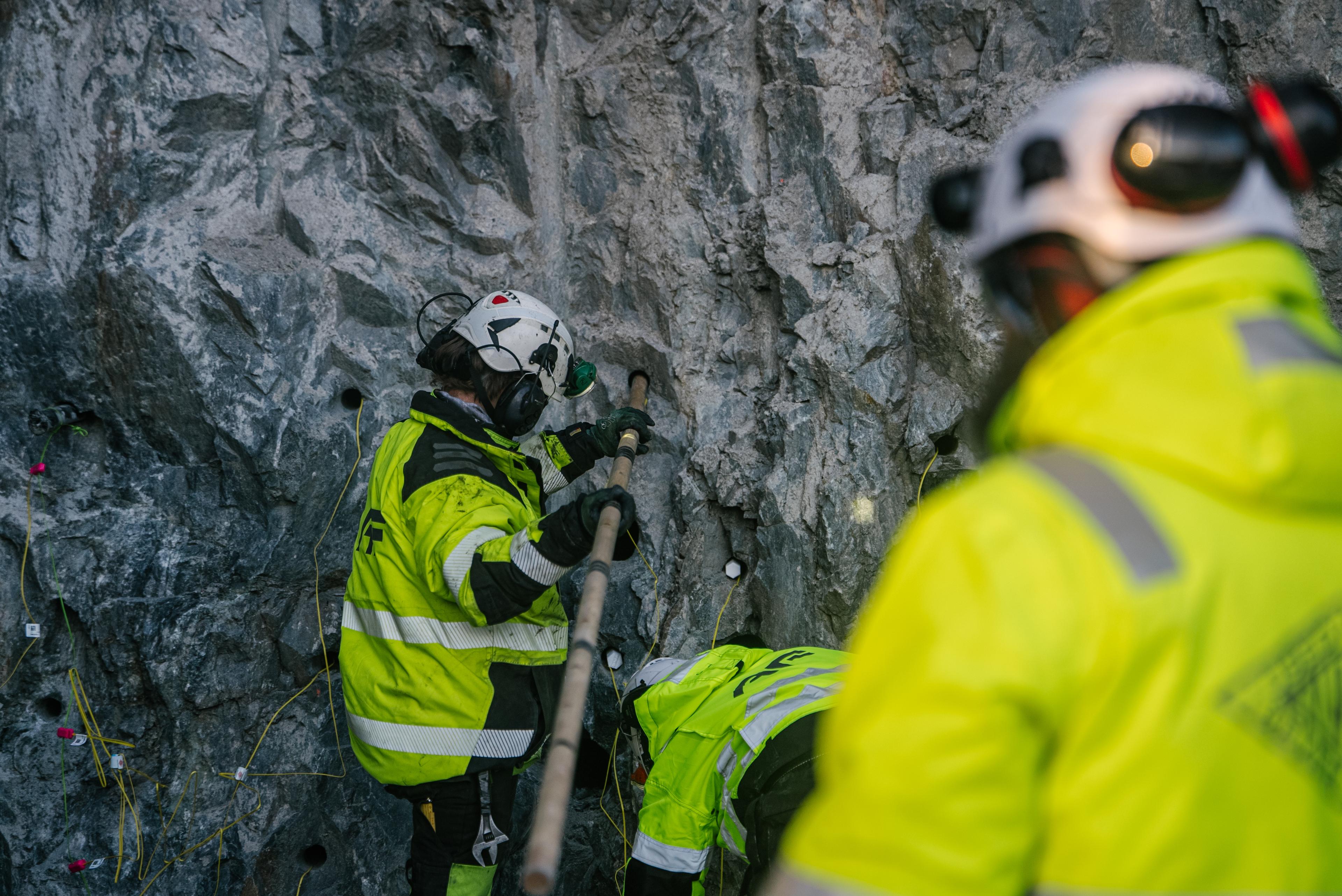 Bildet viser tunnelarbeidere i aksjon ved fjellveggen. En arbeider med hvit hjelm og gul refleksjakke bruker et boreapparat eller stang mot steinoverflaten. En annen arbeider er synlig i forgrunnen. Begge har på seg sikkerhetsutstyr inkludert hjelmer og høysynlighetsklær mens de jobber med fjellsikring eller boring.