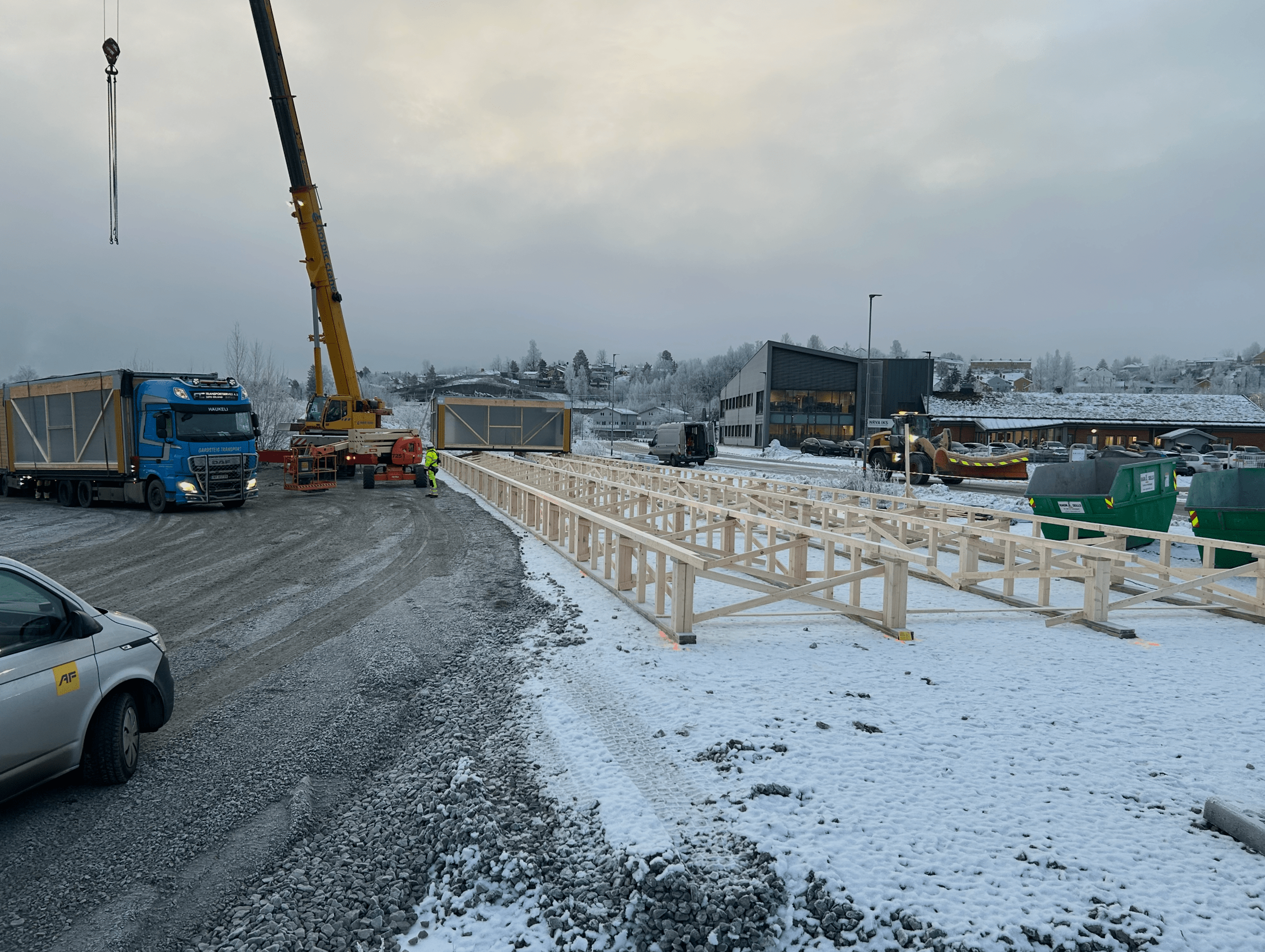 Byggeplass i vinterlandskap med trekonstruksjon, lastebil, byggekran og containere på delvis snødekt område.