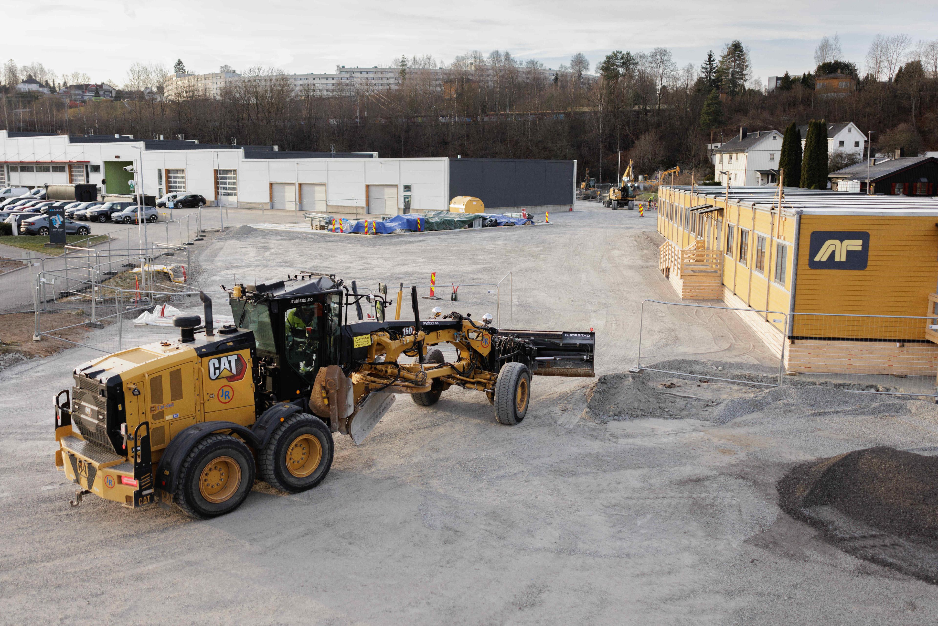 Gul Cat-veihøvel på anleggsområde med gule brakkebygg og industribygninger i bakgrunnen.