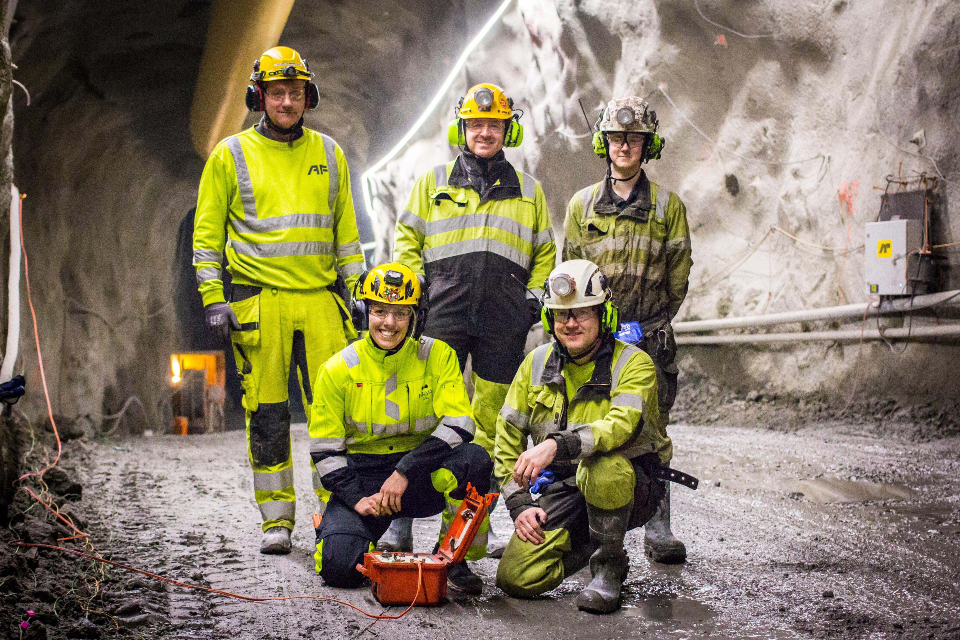 Fem smilende personer i signalgule arbeidsklær står i en tunnel bak et sprengningsapparat.