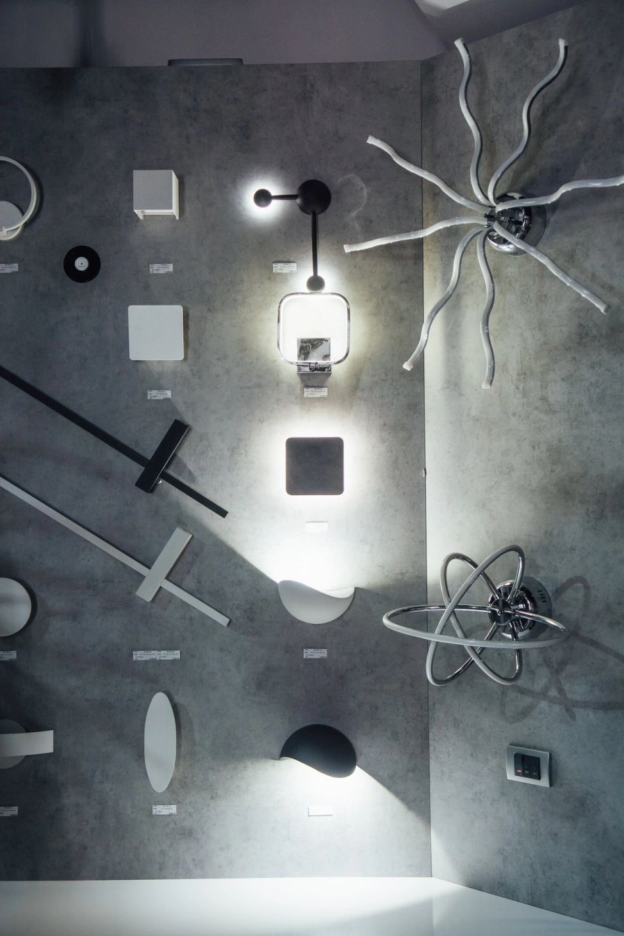 Dve minimalističke zidne LED lampe, bela ovalna i crna zaobljena, upaljene na sivom zidu izložbenog prostora.