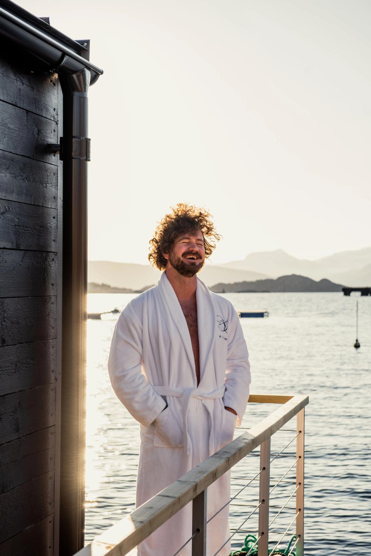 Person looking out a window with a glass of champagne dressed in a bathing towel.