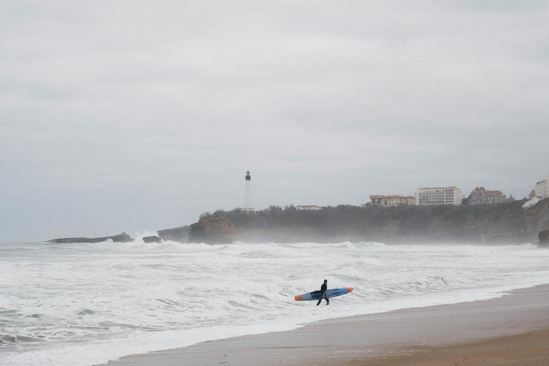 Surfer carrying a board on Biarritz beach with lighthouse and ocean waves in view. Perfect for travel and lifestyle themes.