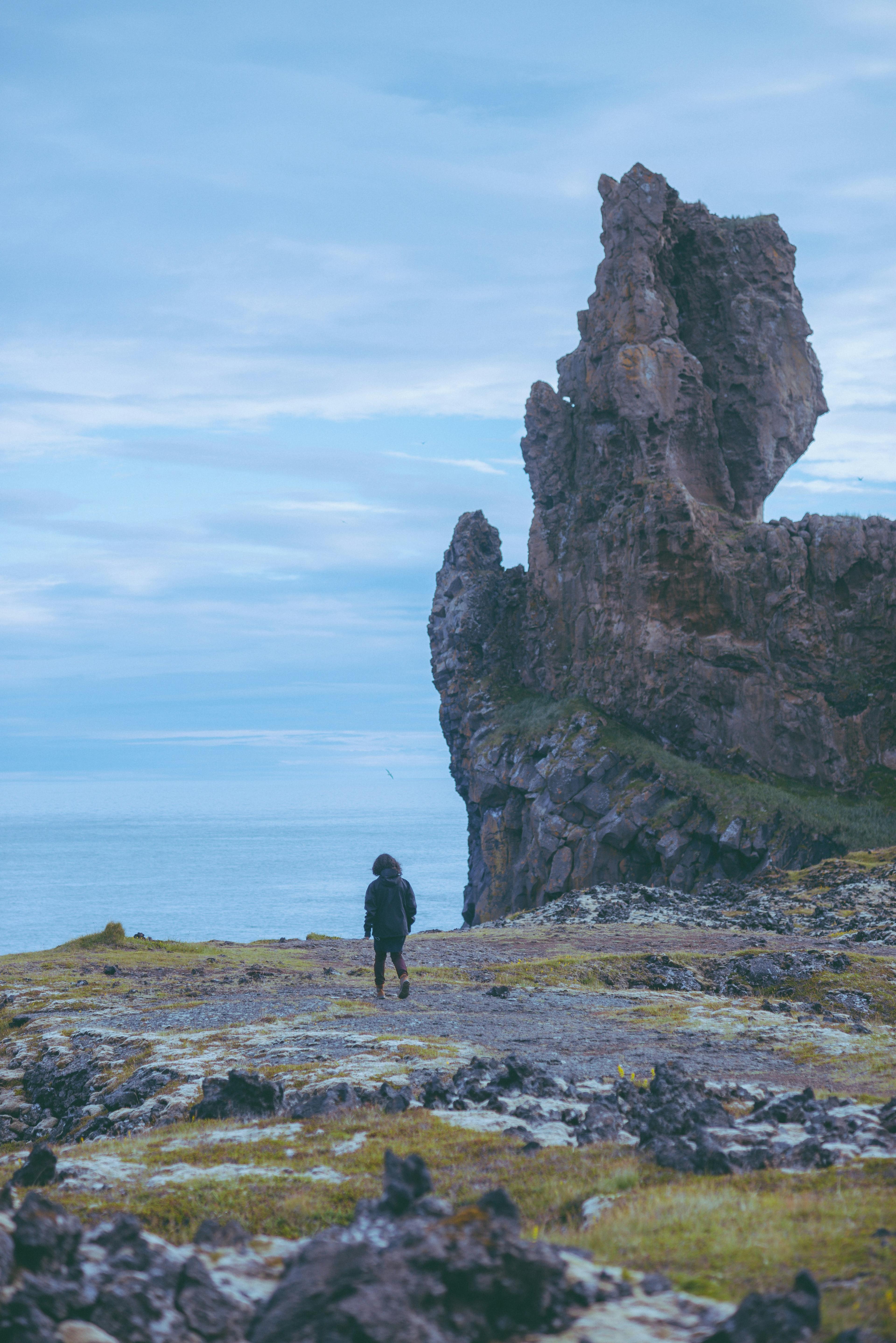 A lone traveler dressed in dark clothing walks toward a towering jagged rock formation on the coast, with the vast ocean and an overcast blue sky in the background