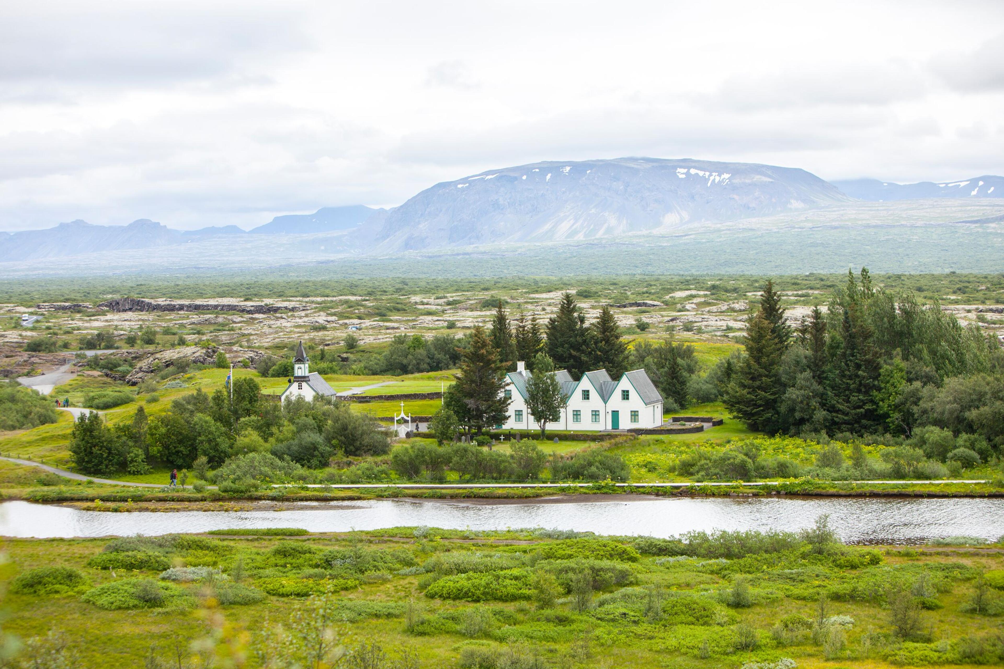 Scenic view of the Thingvellir National Park in the Golden Circle, Iceland, featuring a cluster of traditional white houses with green roofs, a church, and a reflective pond surrounded by lush greenery with distant mountains under a cloudy sky
