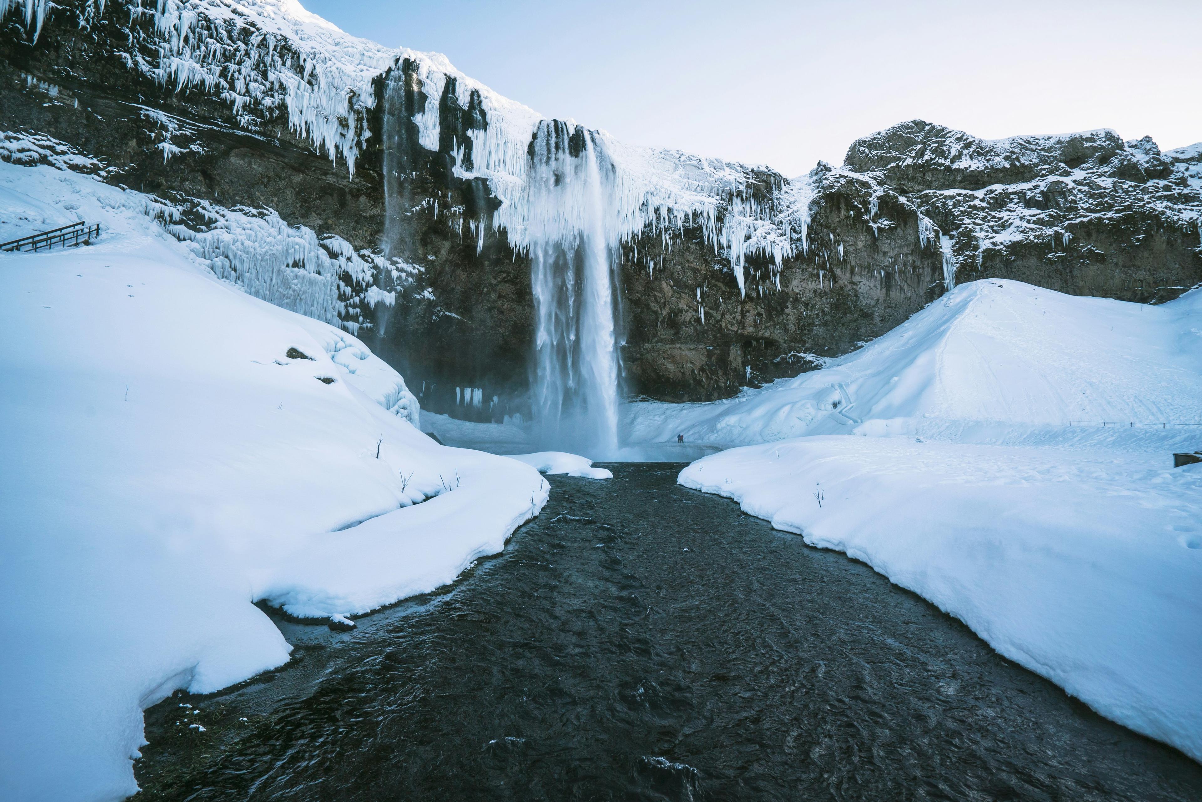 Seljalandsfoss waterfall cascades over frozen cliffs into a snow-lined river under a bright, clear winter sky.