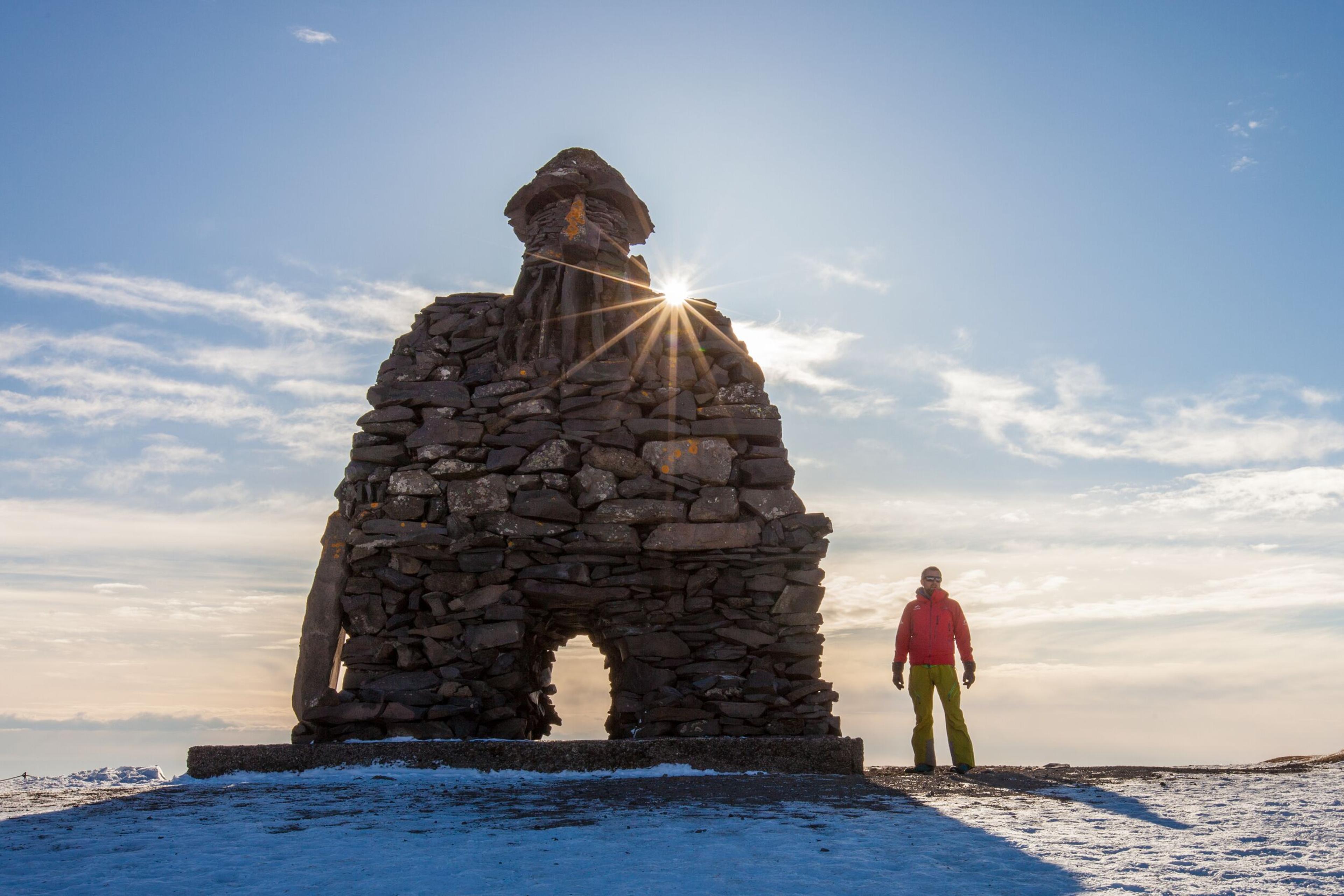 A large stone sculpture resembling a traditional Icelandic guardian figure stands on a snowy landscape, with the sun creating a starburst effect through its archway. A person in red stands nearby, adding scale to the scene.