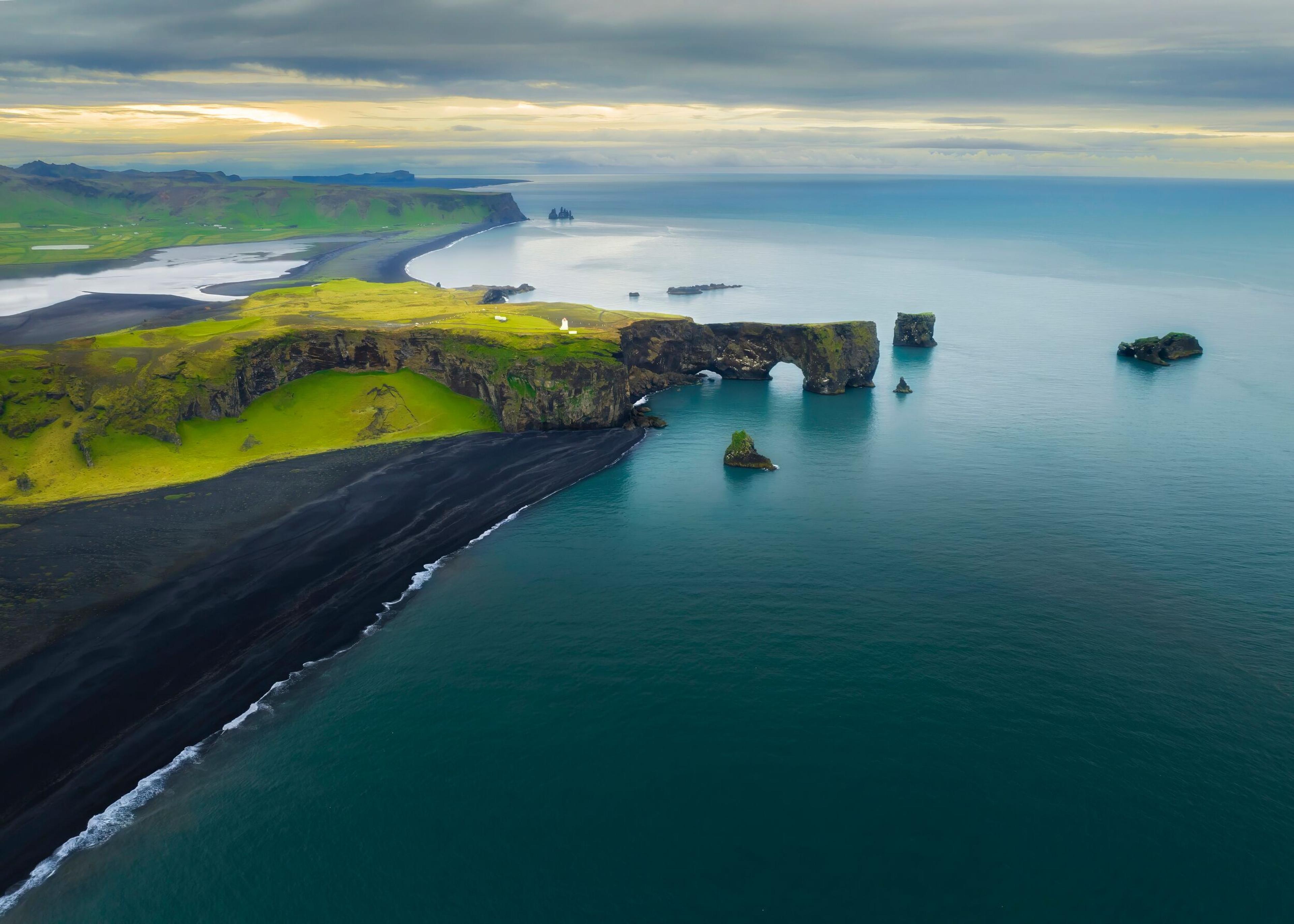 Aerial view of a dramatic coastline with black sand beaches, grassy cliffs, and rock formations jutting out from the sea under a cloudy sky.