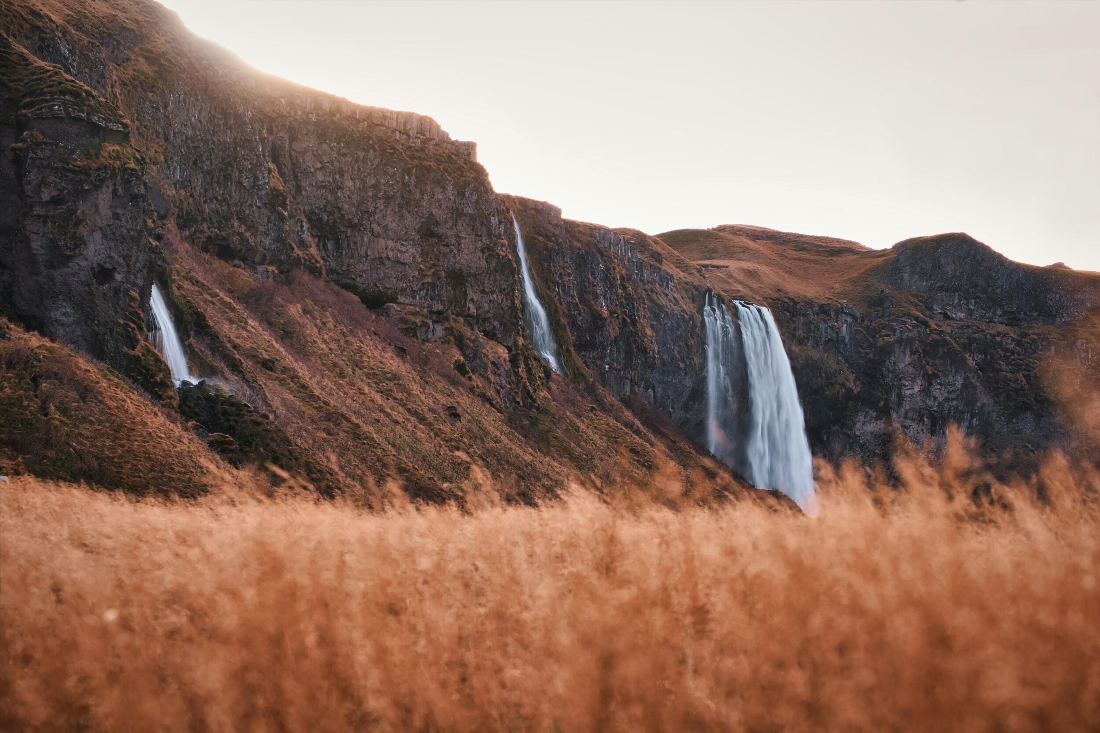 Distant view of a waterfall cascading down rocky cliffs surrounded by golden grass on Iceland's South Coast.