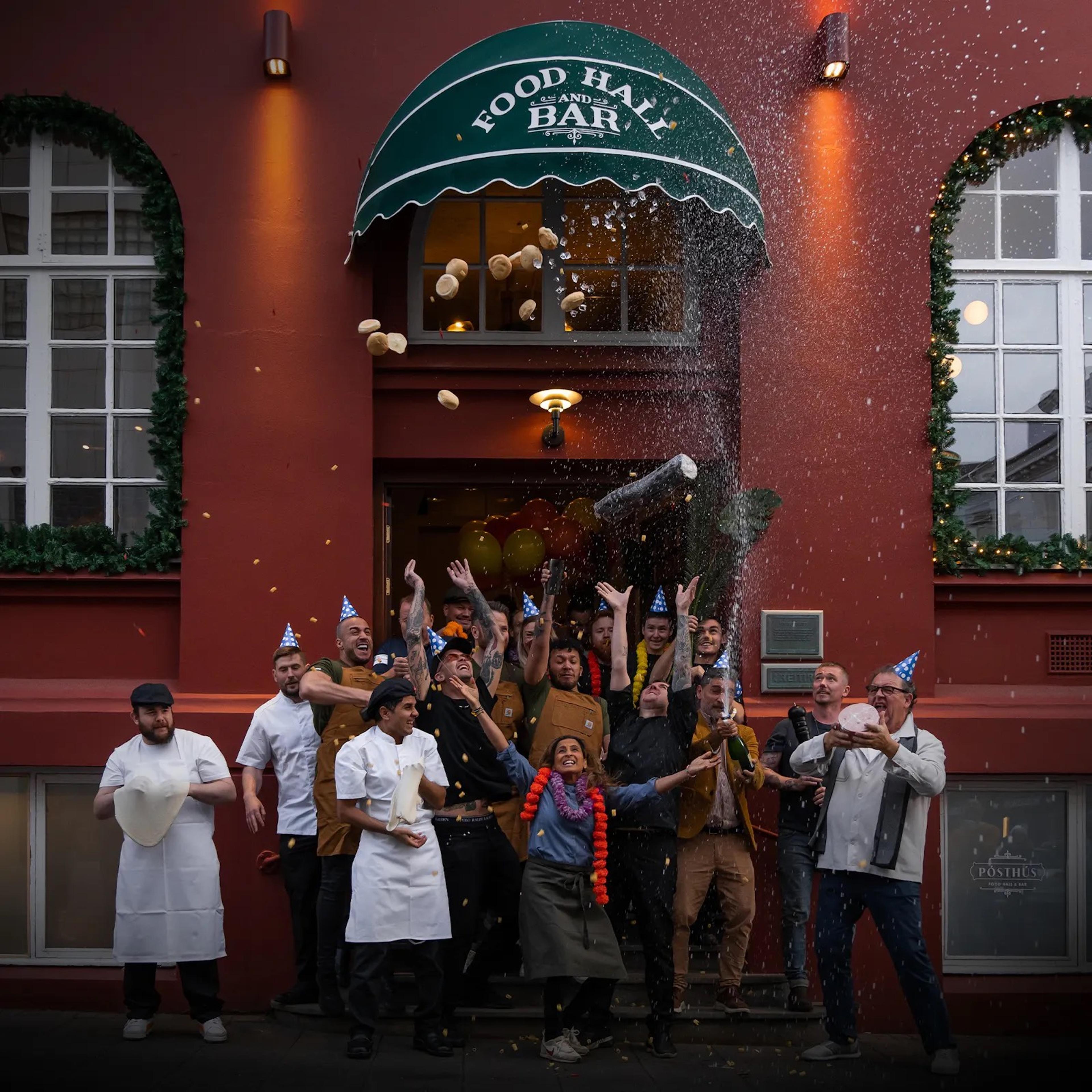 Celebratory scene outside Posthús Food Hall, with chefs and staff wearing party hats, throwing bread, and popping champagne under a green awning.