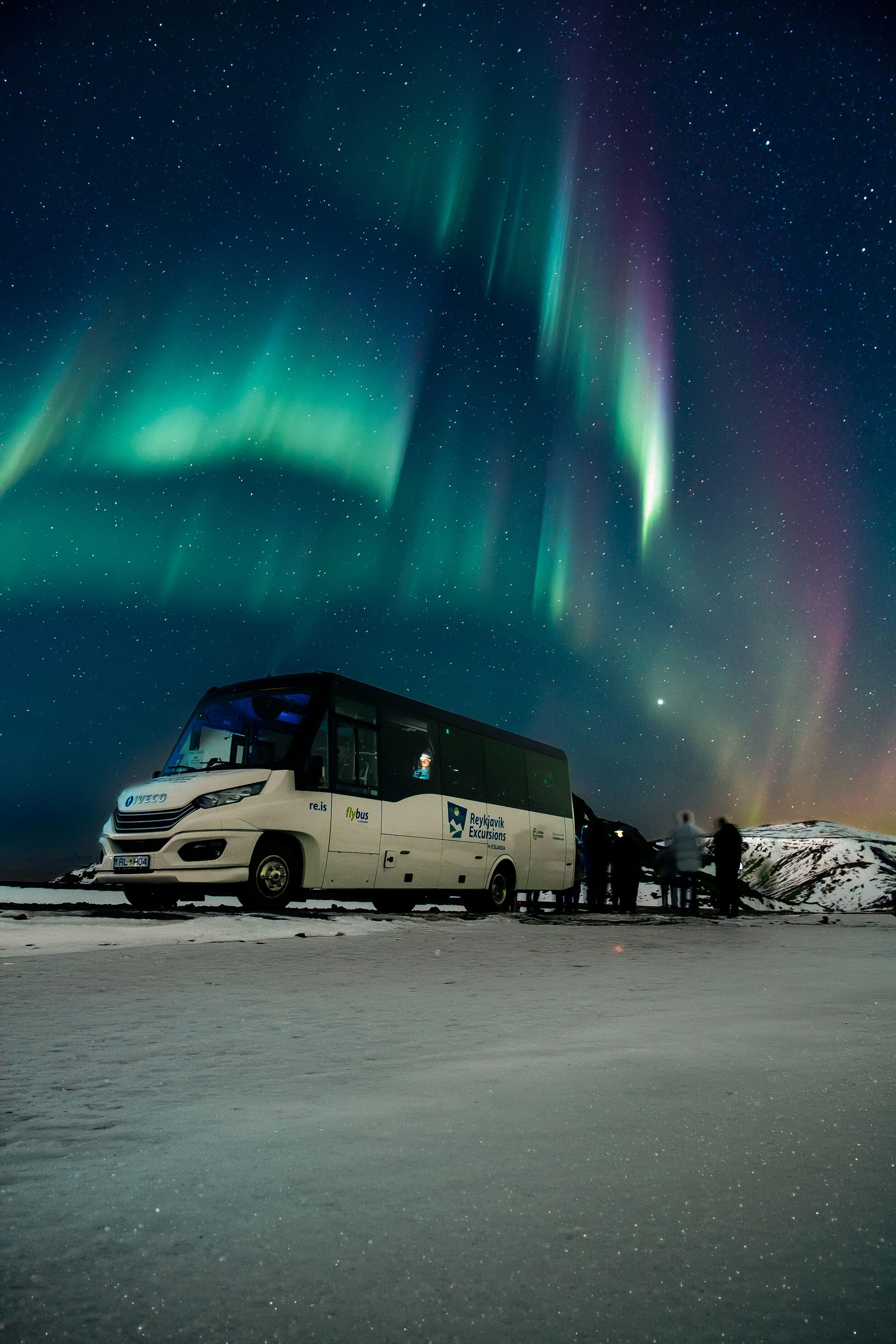 A white tour bus parked on a snowy field under a starry night sky, illuminated by vibrant green and purple Northern Lights, with people watching.