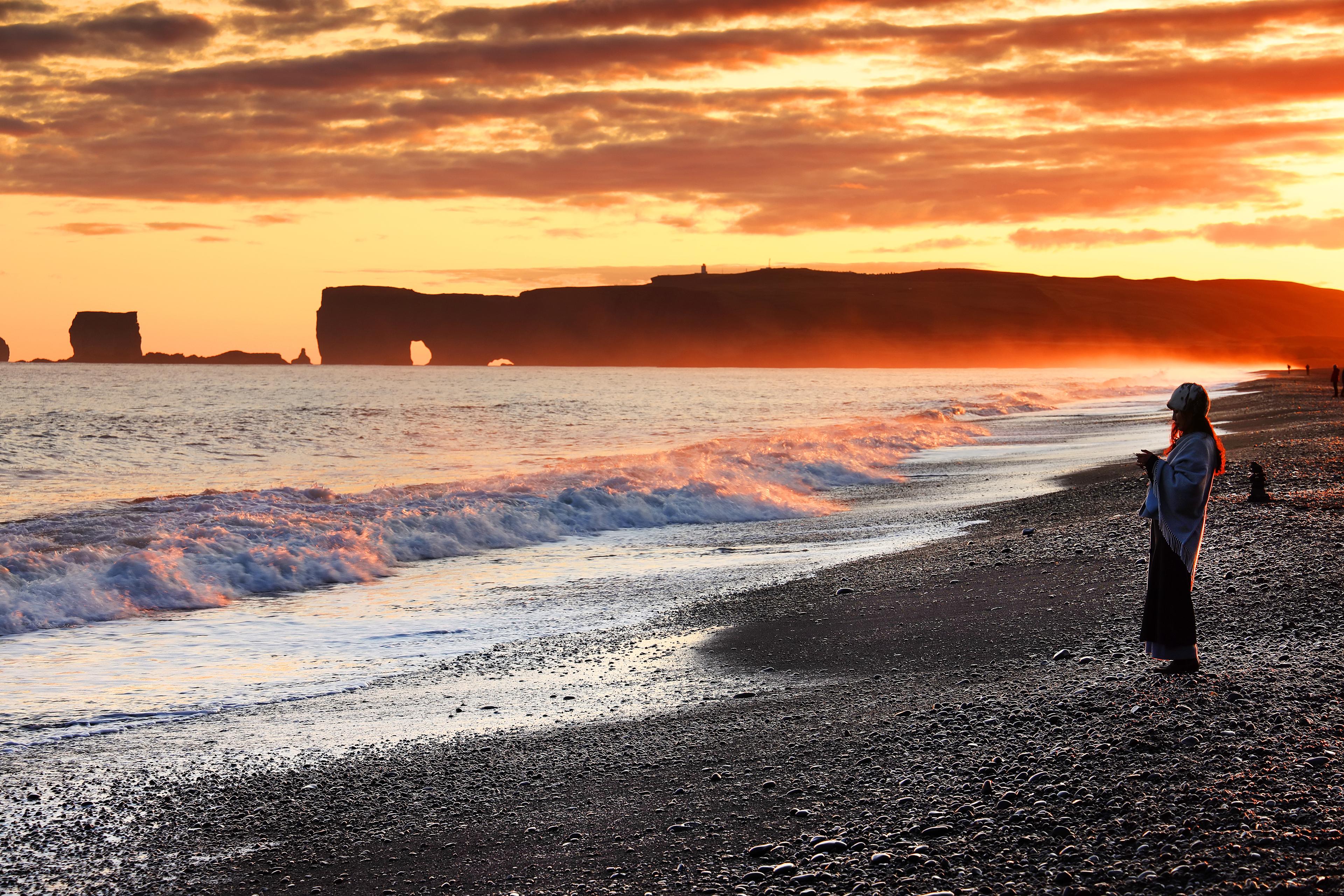 A person on the Reynisfjara Beach during sunset. Dyrhólaey appears on the background.