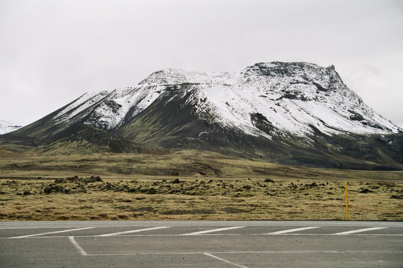 A snow-capped mountain towers over a vast, dry landscape with an asphalt road in the foreground under an overcast sky.